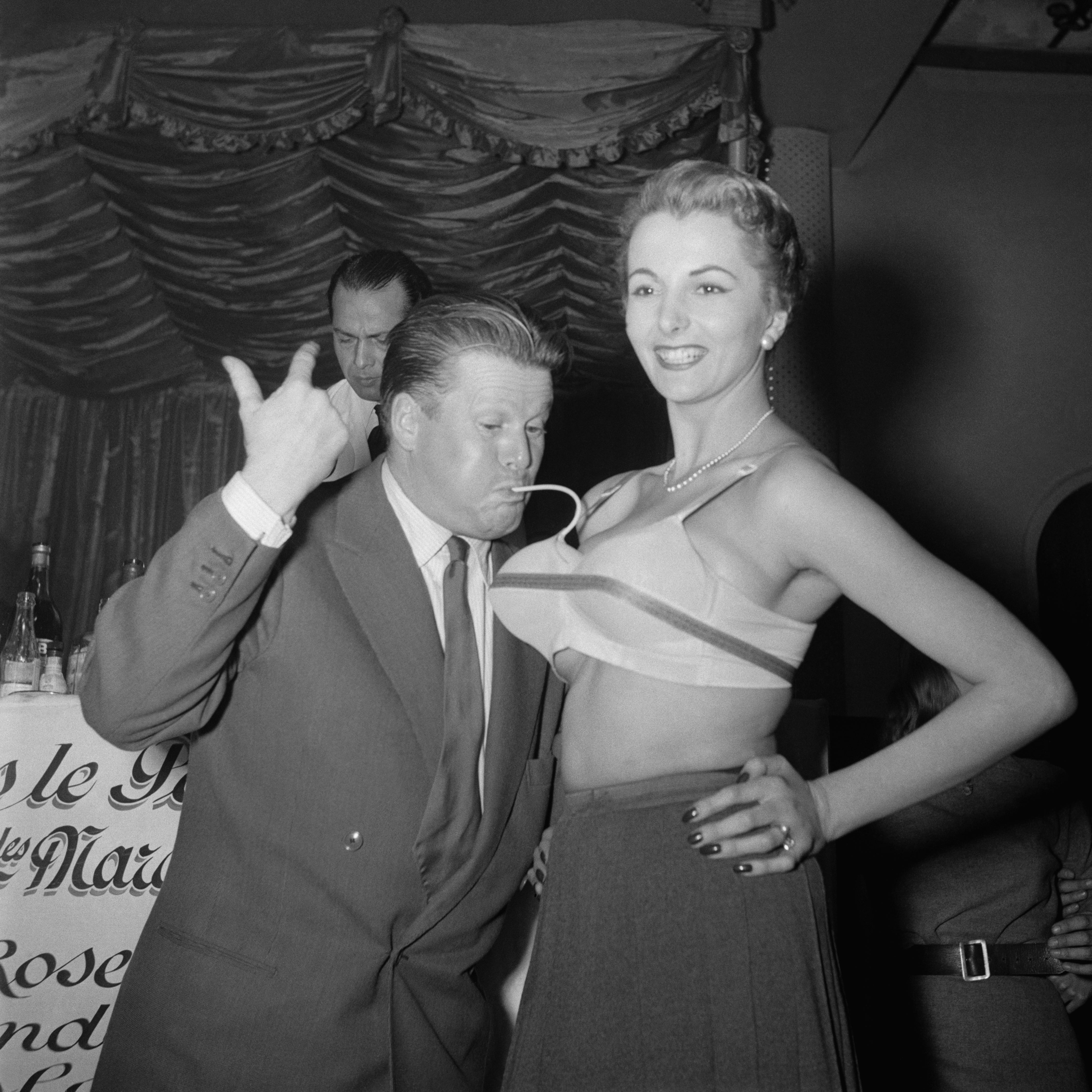Jacques Angelvin playfully interacts with a woman in an inflatable bra at an event, with another man in the background