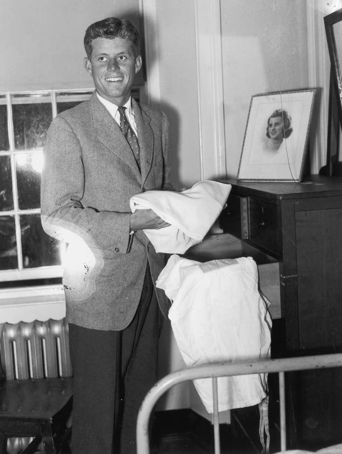 John F. Kennedy smiles while folding laundry in a room with a framed photo on a dresser