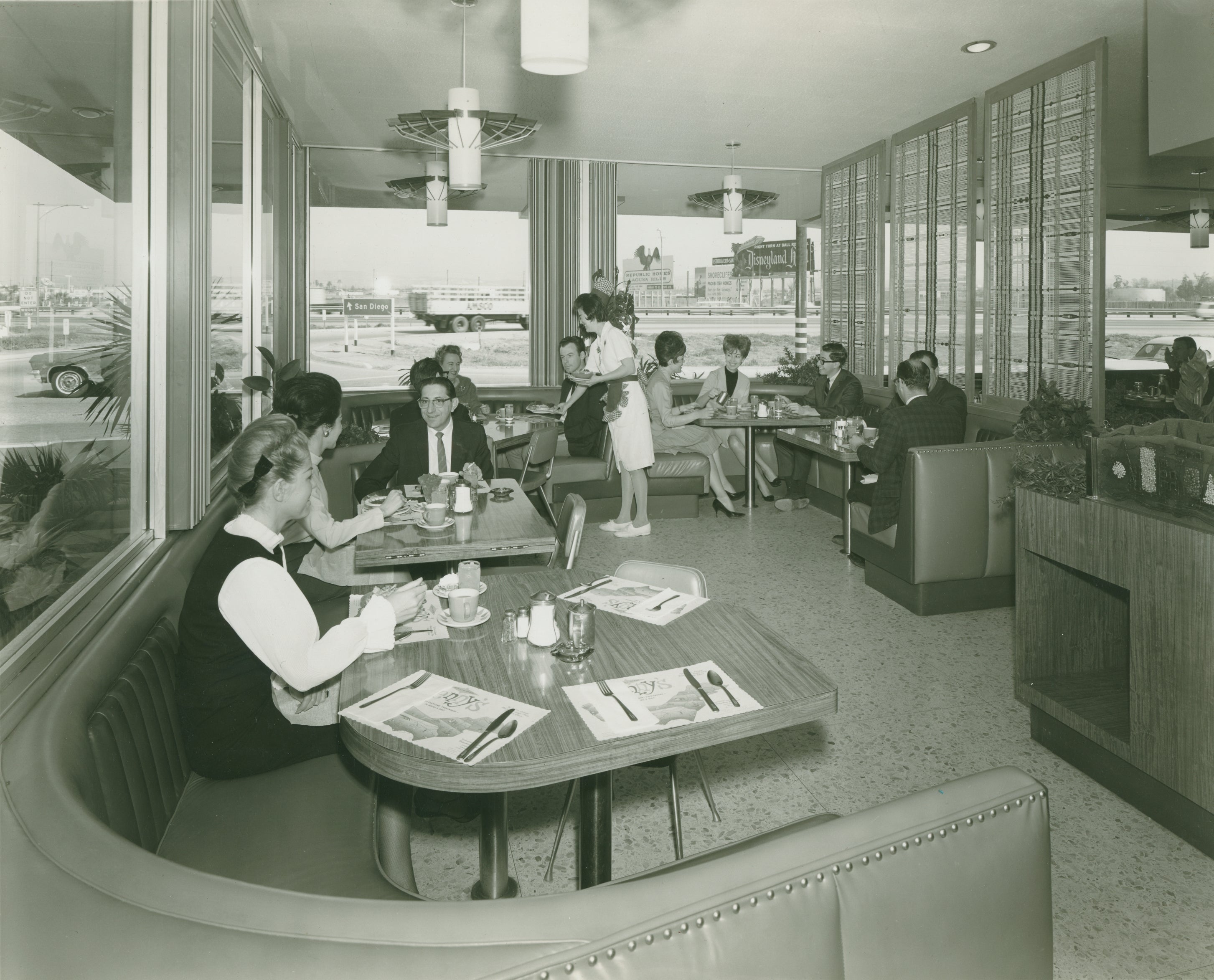 A vintage diner scene with patrons seated at tables, a waitress serving food, and large windows showing a view outside