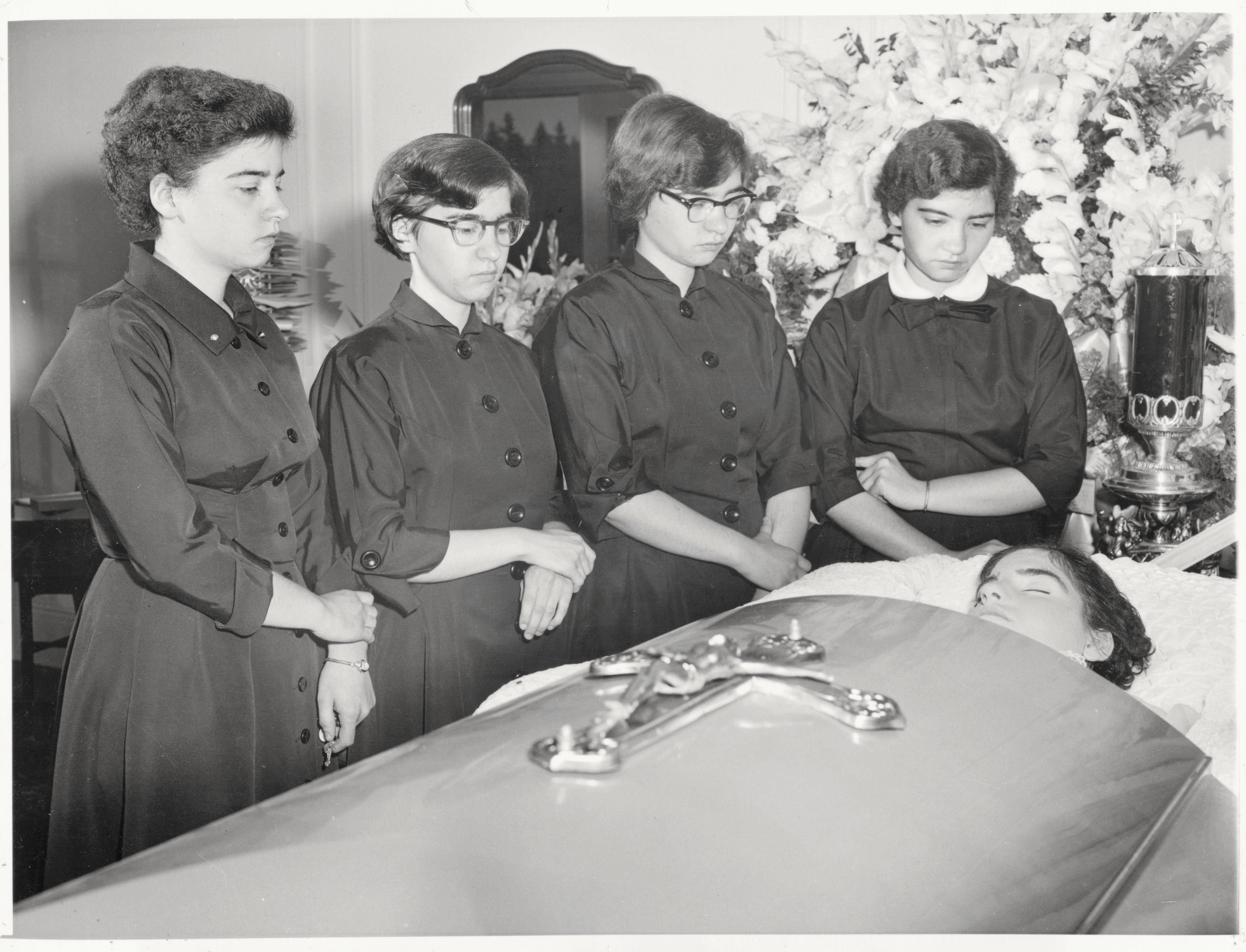 Four women stand solemnly by a casket, paying respects at a funeral. They wear similar formal dresses