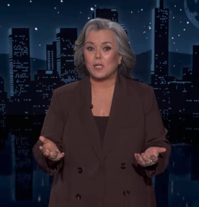 Person in a suit speaking on a nighttime talk show set, cityscape backdrop visible