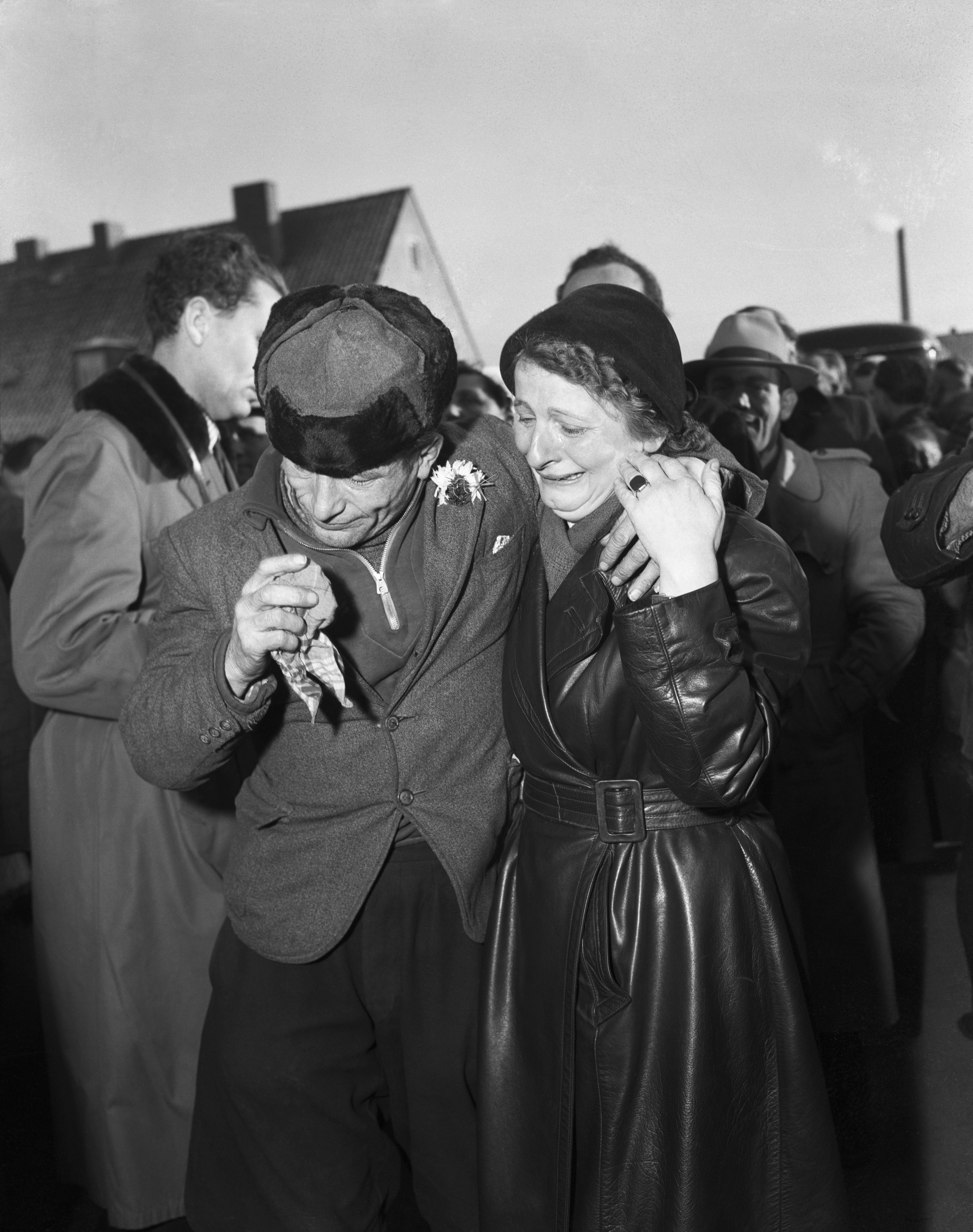 A man in a cap hugs a crying woman in a belted coat as they walk in a crowded outdoor setting