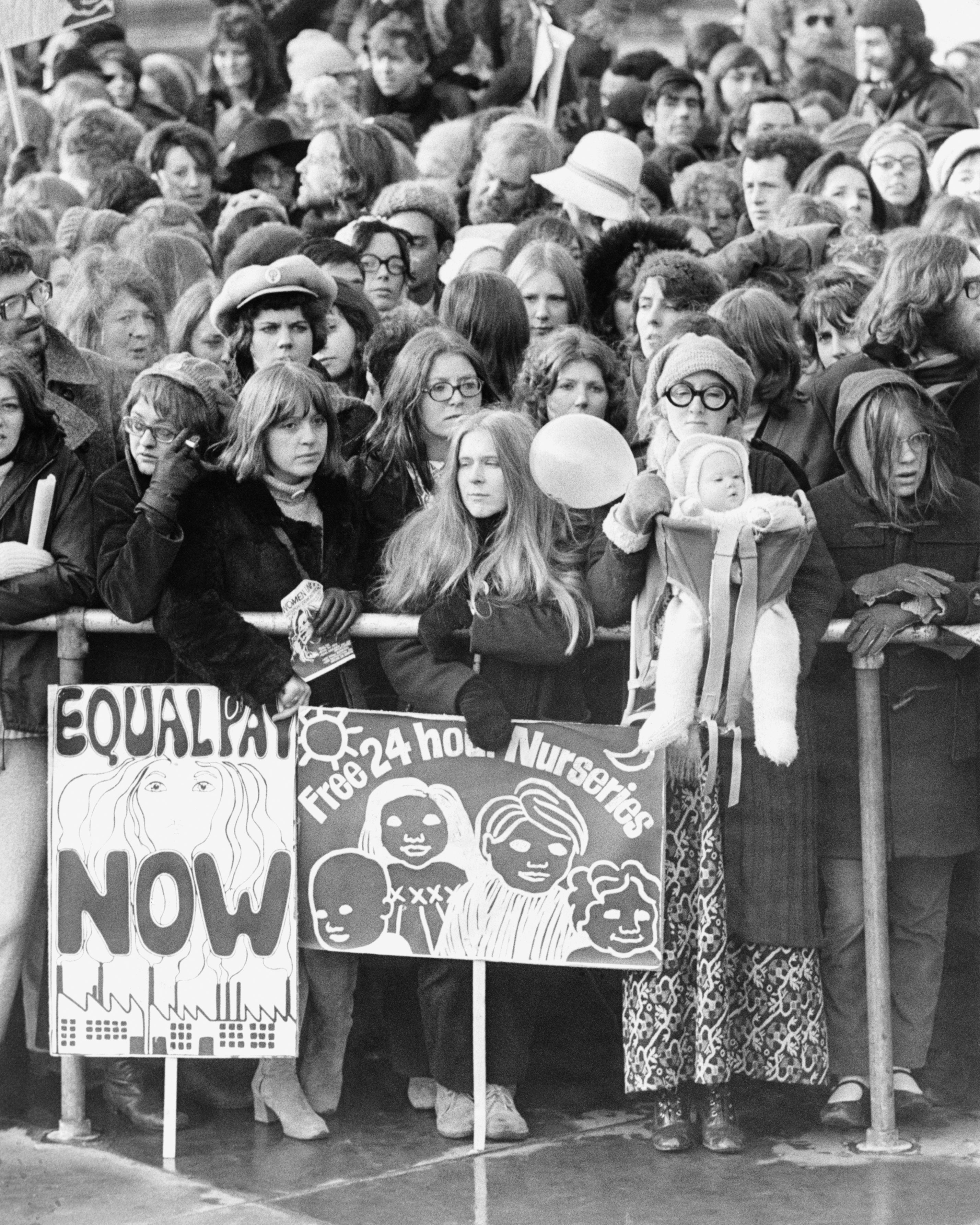 A large crowd of women at a protest holding signs that read "Equality Now" and "Free 24 hour Nurseries" with illustrations