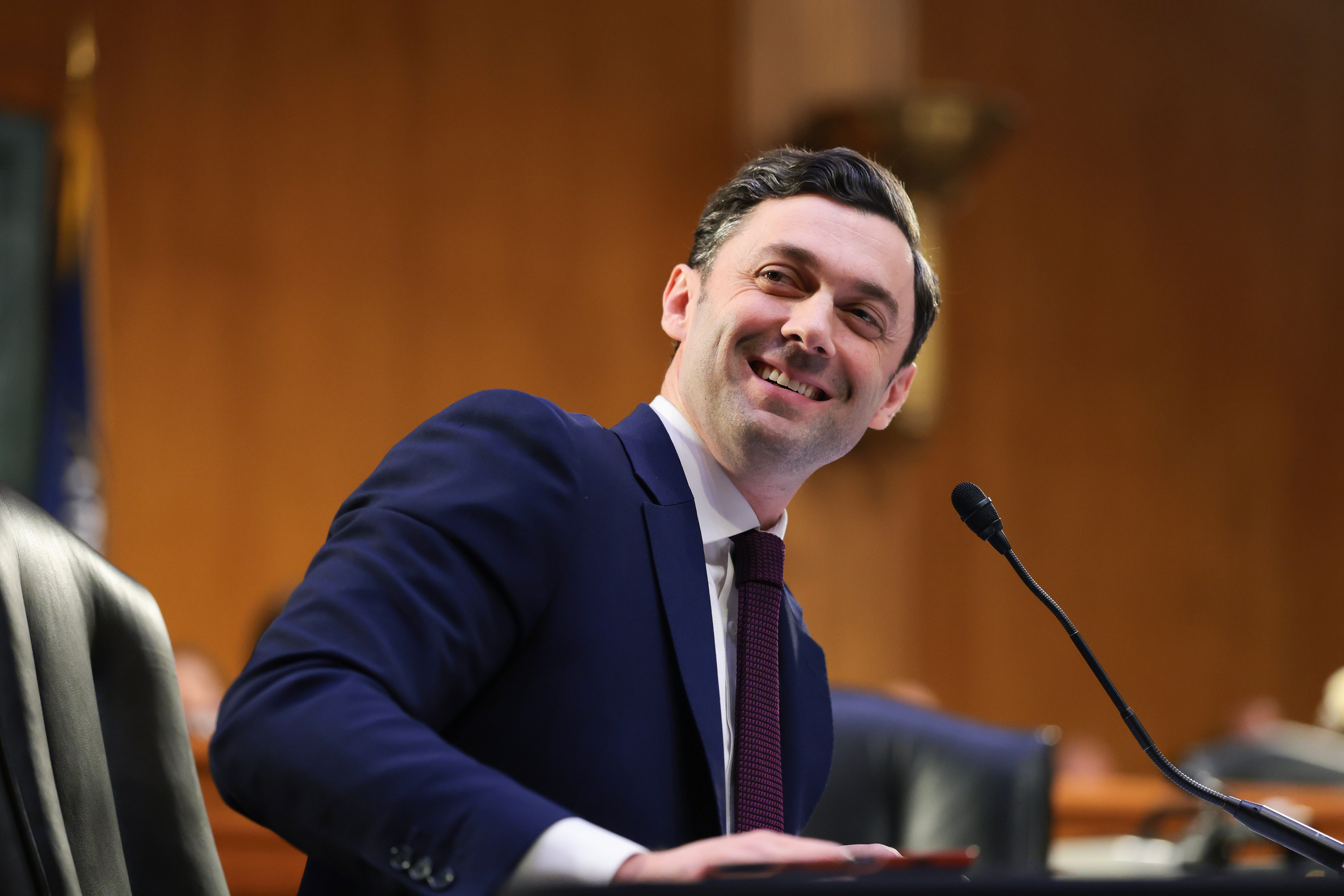 Person in a suit, sitting at a desk, smiling and turning slightly to the side. Office setting