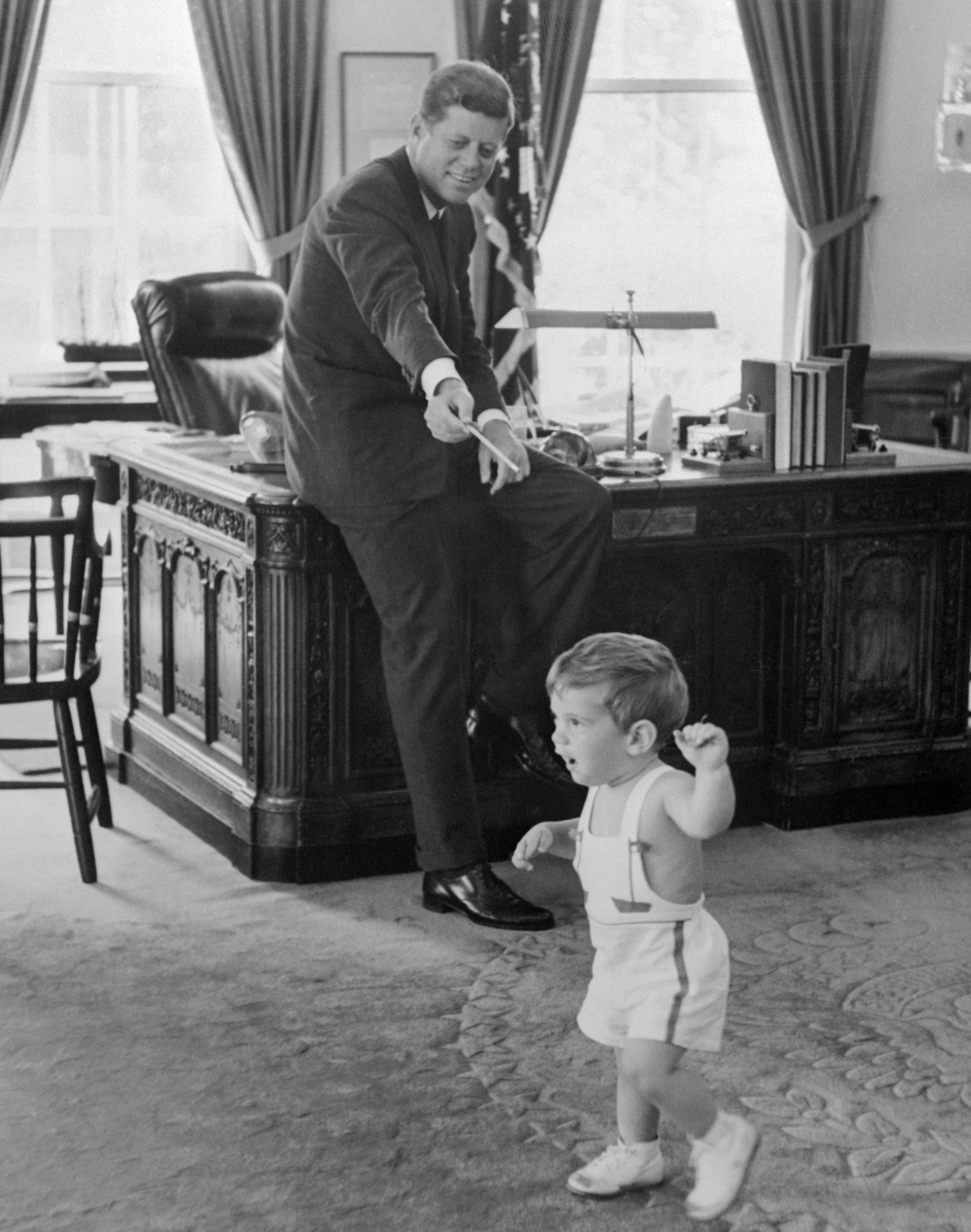 John F. Kennedy playfully gestures from his desk toward a toddler, JFK Jr., walking in an office