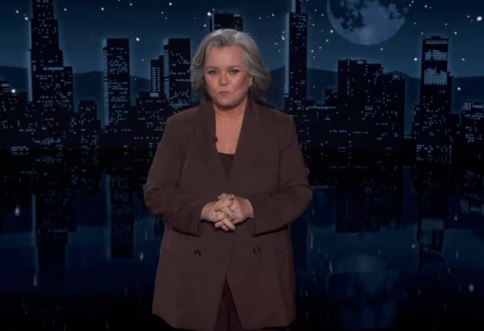 Person stands confidently on stage in front of city skyline backdrop, wearing a tailored suit