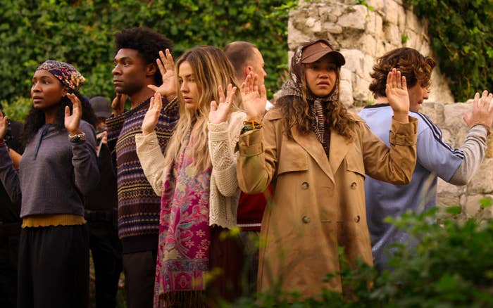 Group of people stand with hands raised, wearing varied casual and layered clothing, in an outdoor setting with stone walls and greenery