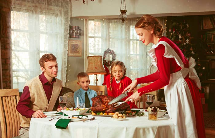 Family at a dining table enjoying a festive meal. A woman is carving a roast, and two children look on joyfully
