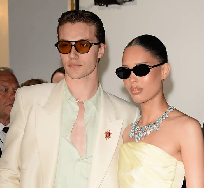 A stylish couple wearing sunglasses and elegant attire pose together at an event