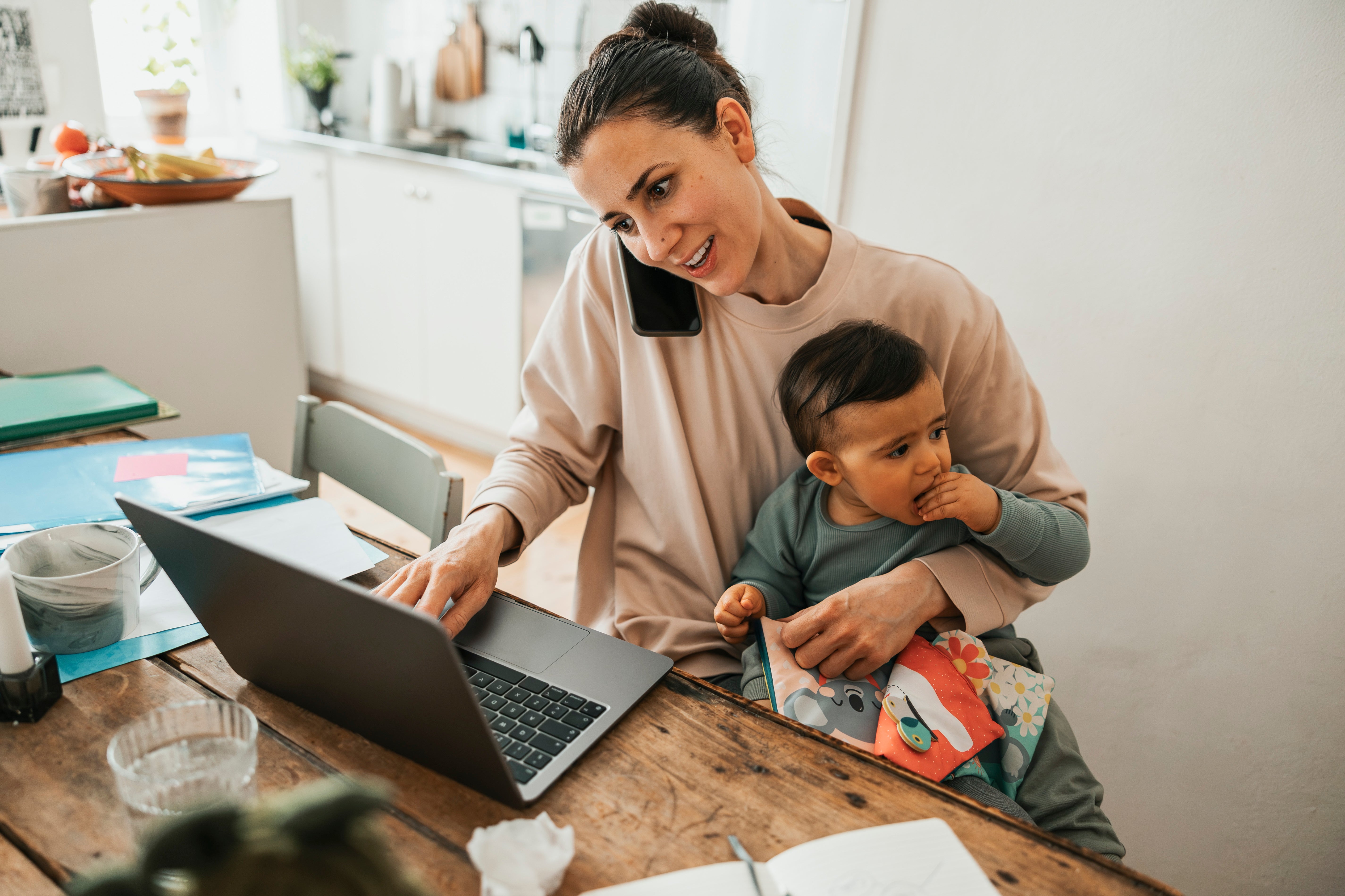  on phone, using laptop, holding a baby who looks curious and is sitting on her lap at a kitchen table