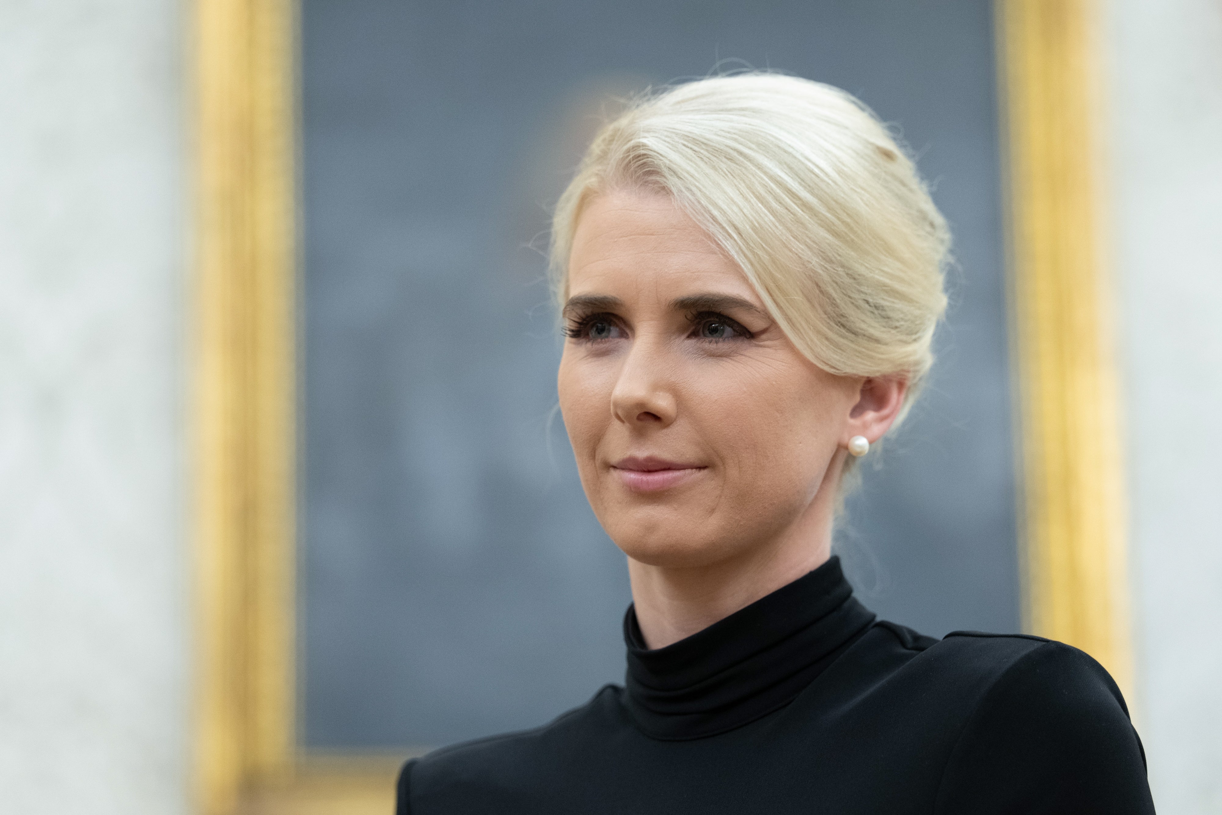En person med blondt hår iført en svart turtleneck står i et rom med et innrammet bilde i bakgrunnen