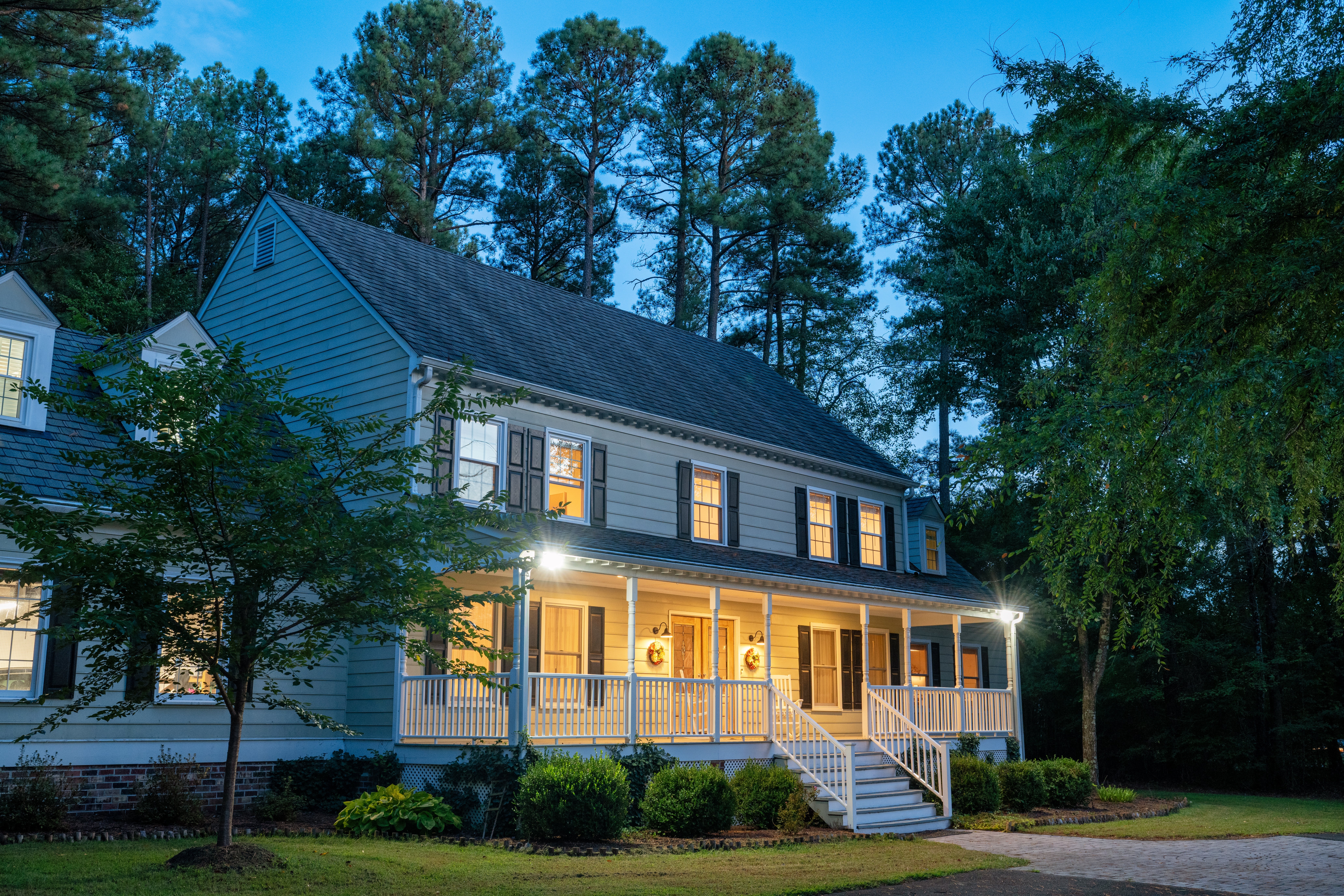 A charming two-story house with a porch, lit warmly in the evening, surrounded by tall trees