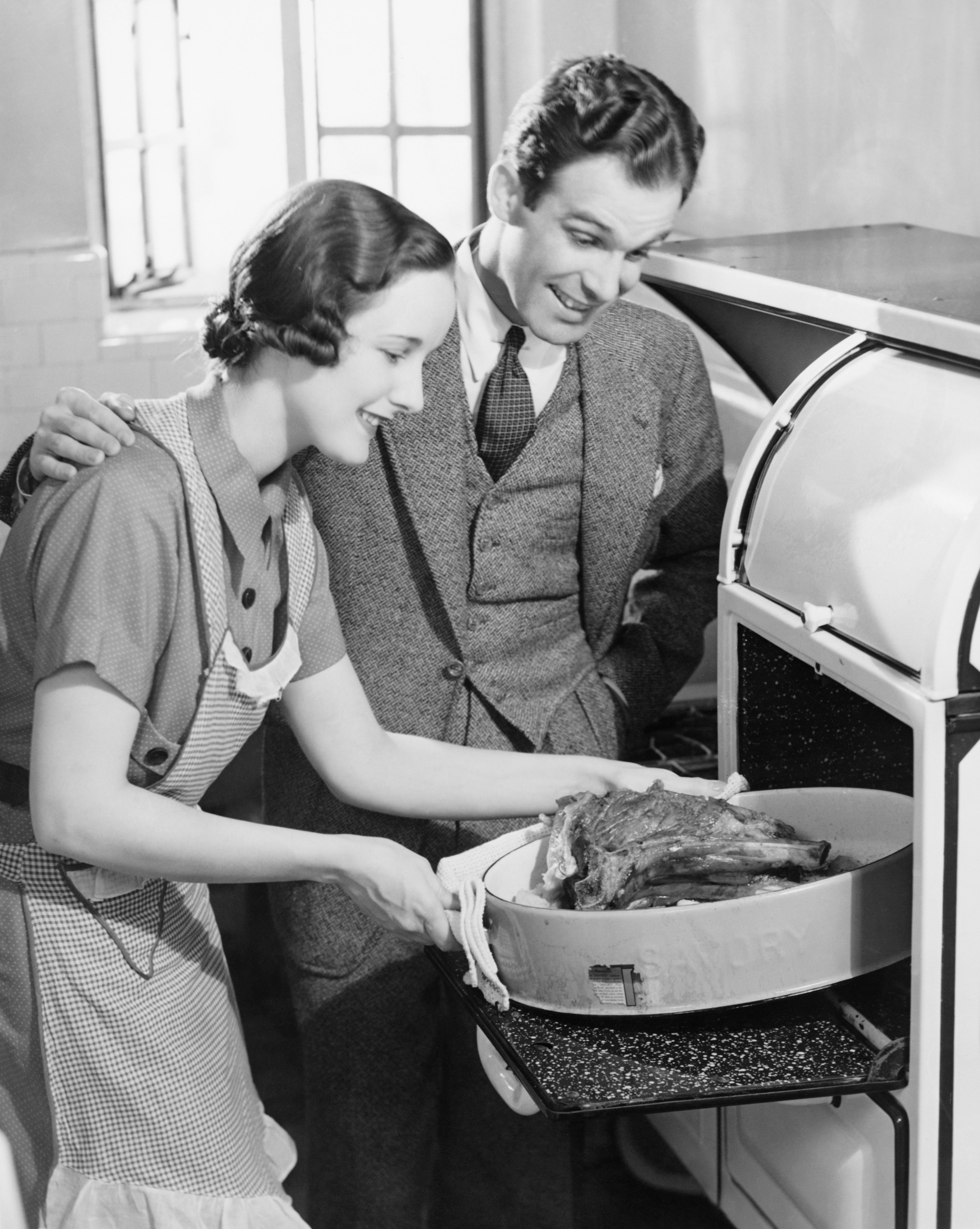 A smiling couple in vintage attire takes a roast out of an oven, showcasing 1930s kitchen life