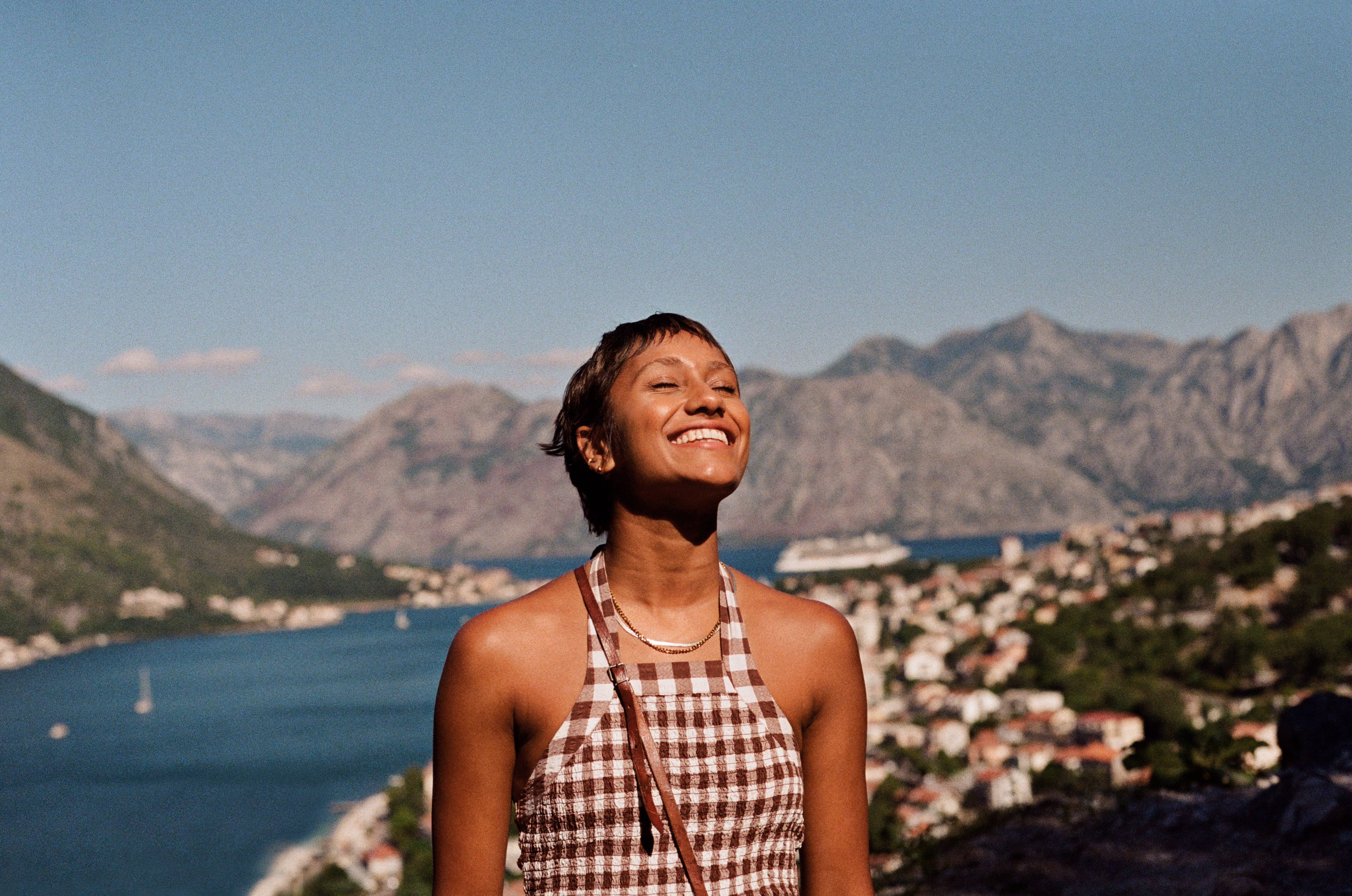 Person smiling in front of a scenic mountain and lake view, wearing a checkered halter dress