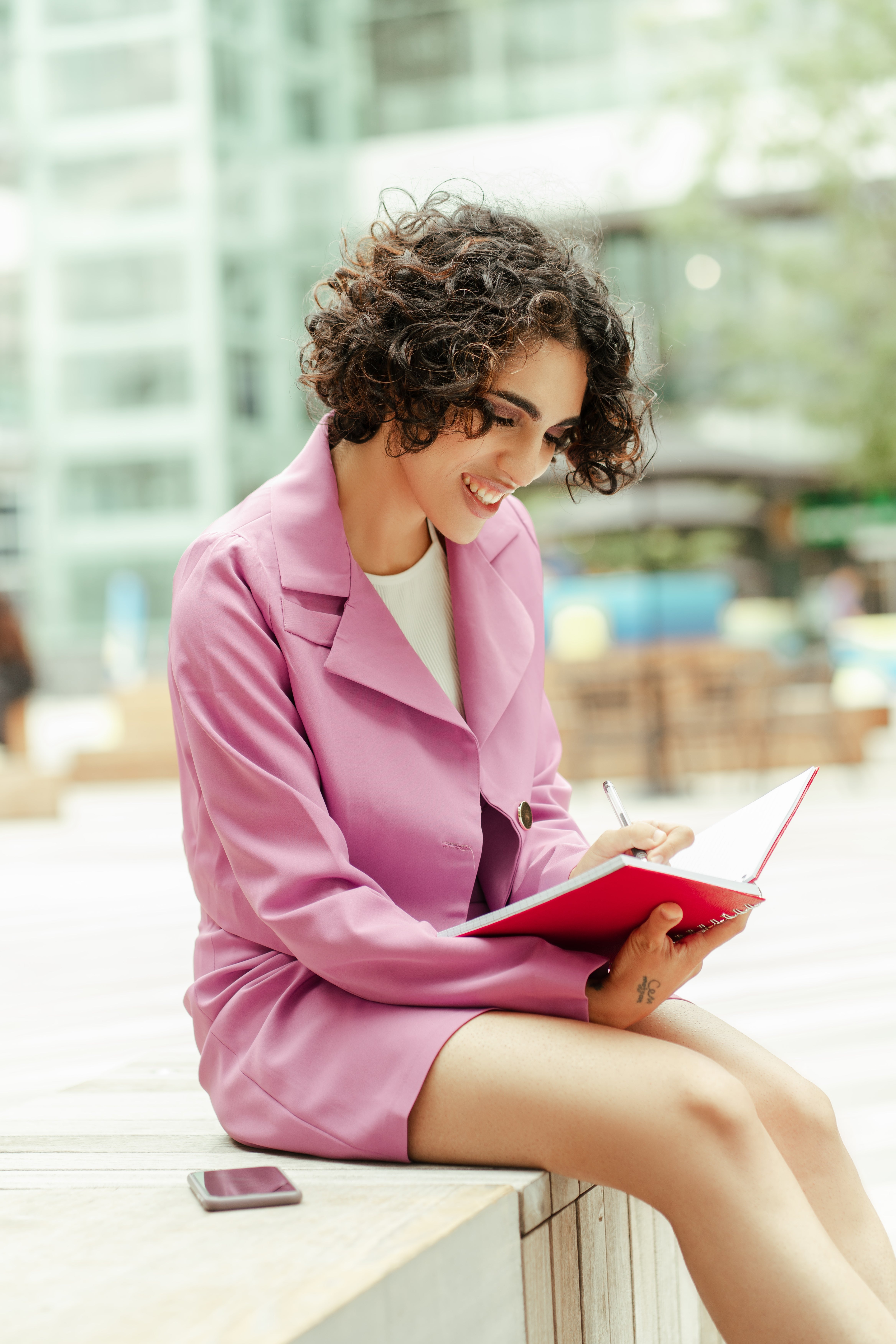 Person sitting outdoors, smiling, and writing in a notebook. They wear a stylish blazer dress and have curly hair. A smartphone is placed nearby