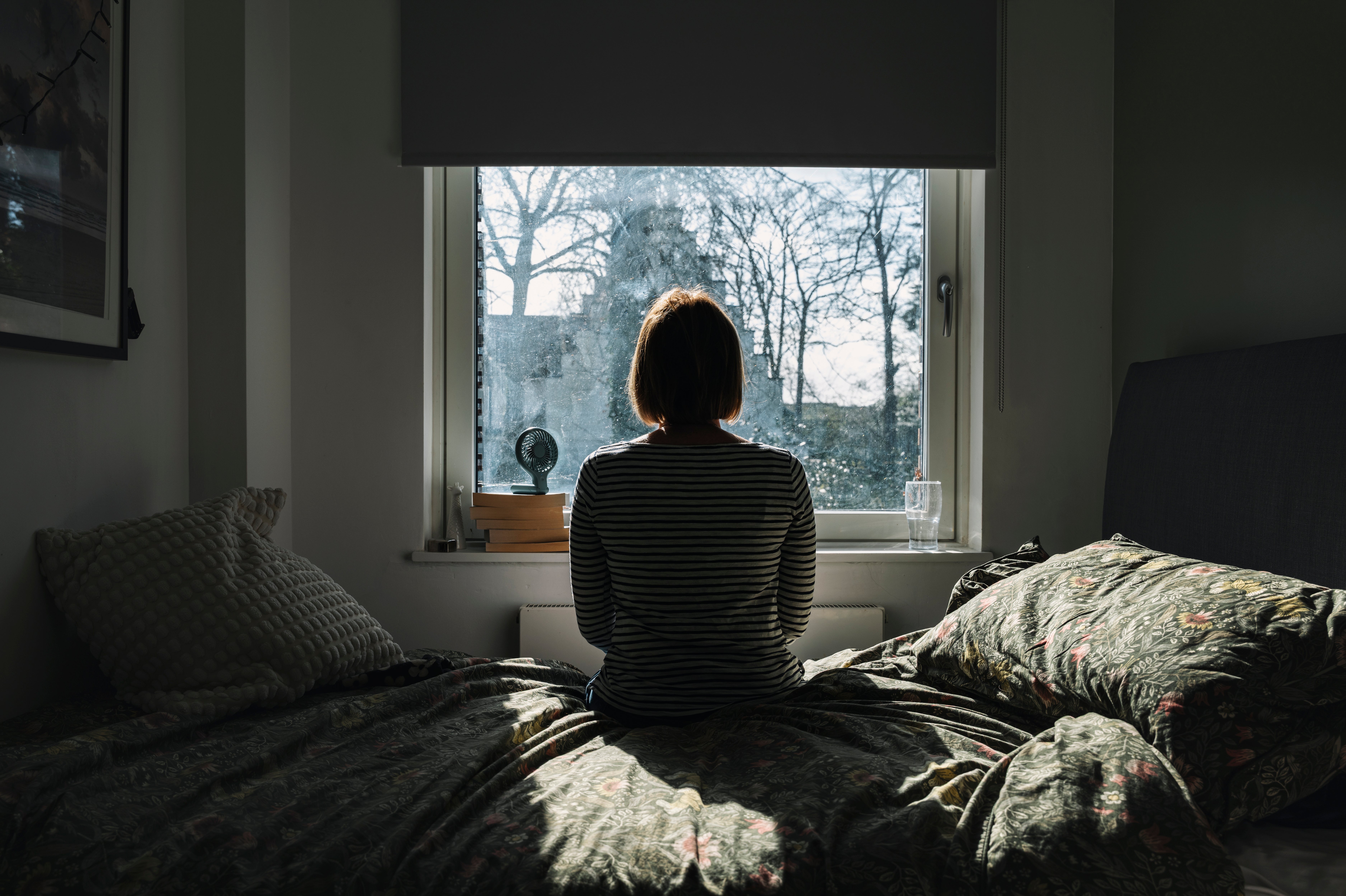 Person sitting on a bed, gazing out a window at a sunny view, creating a reflective and calm atmosphere in the room