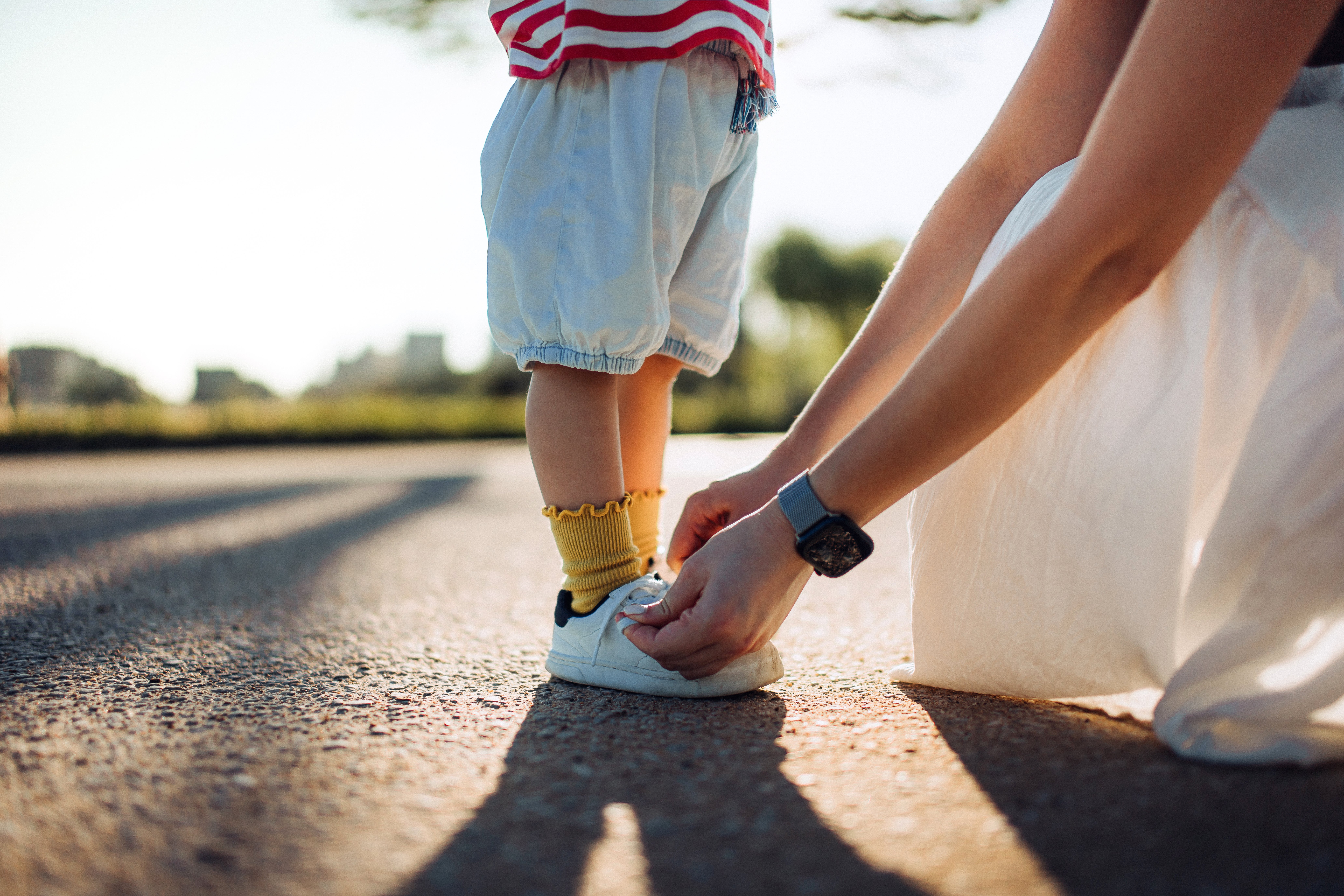 Adult tying a child's shoe on a sunny day, focusing on the child's legs and feet