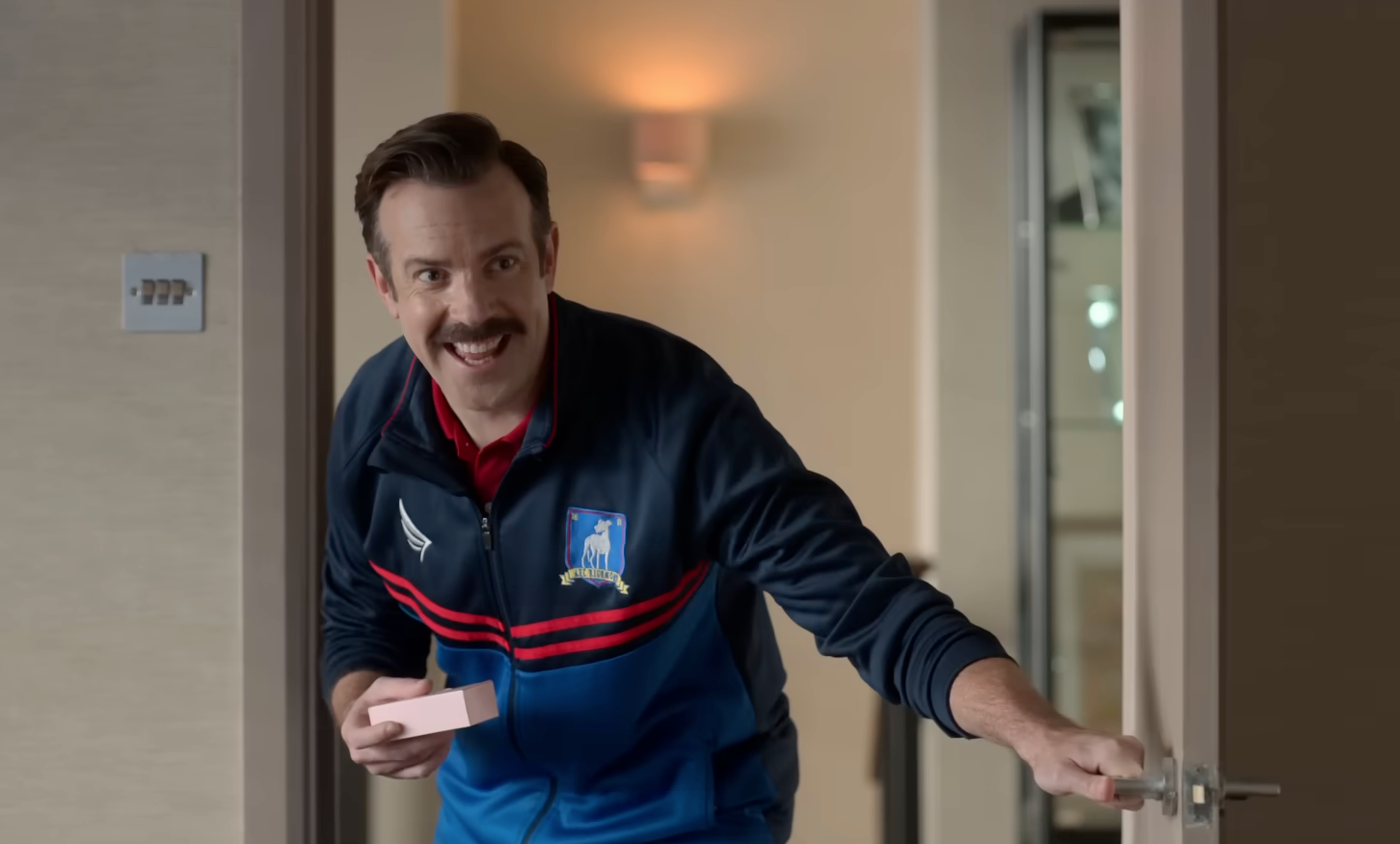 A person with a mustache wearing a sporty jacket opens a door with a smile, holding a small box