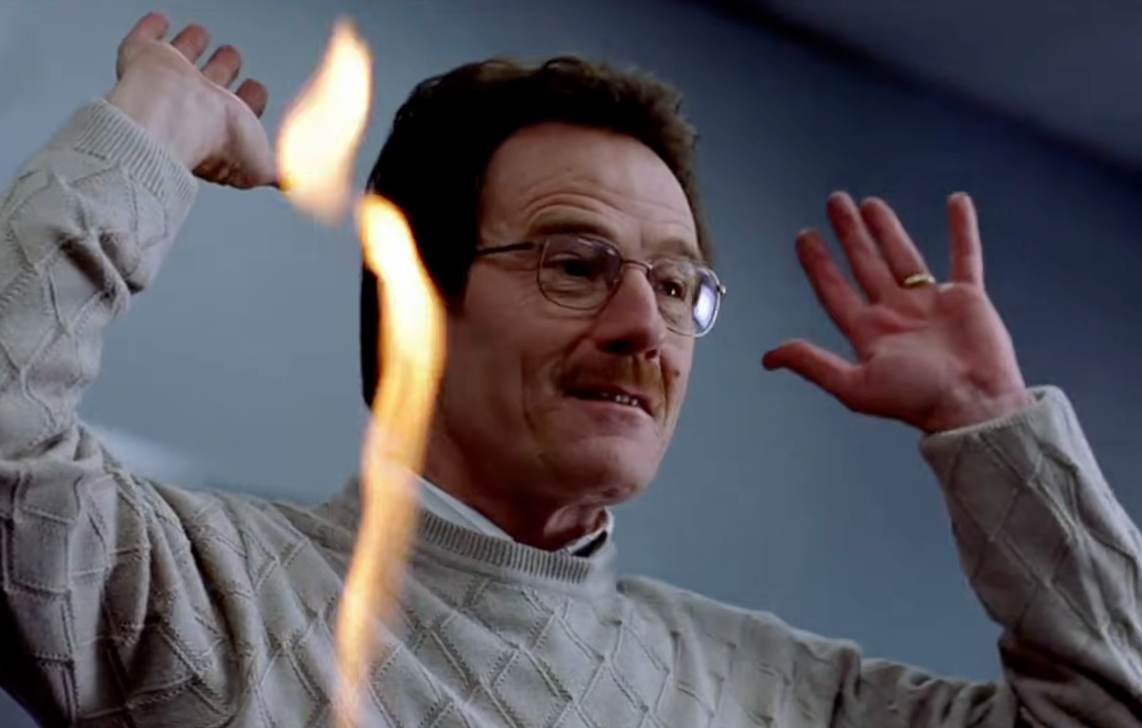A man in glasses and a patterned sweater holds his hands up in a tense scene with a flame in the foreground