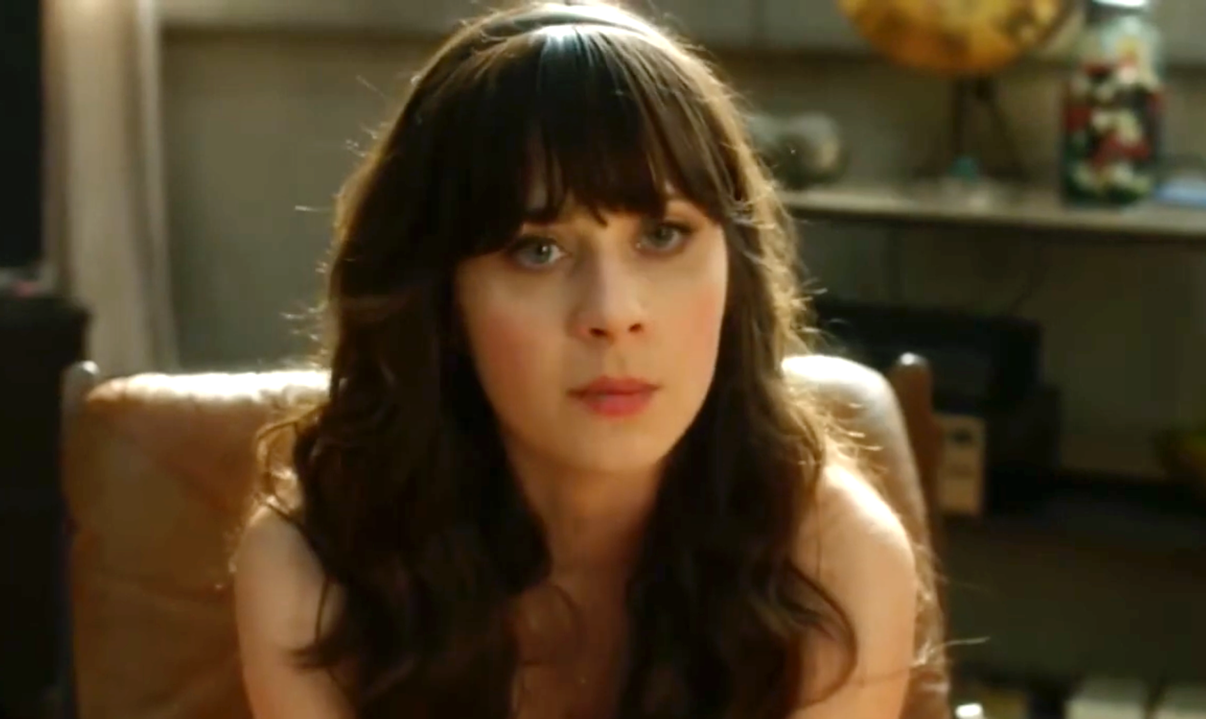 Person with long dark hair and bangs sits indoors, looking forward with a thoughtful expression. Background features shelves and decor
