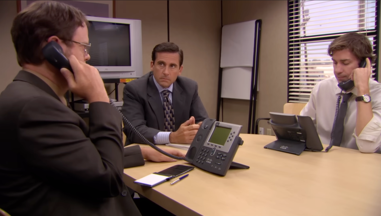 Three office workers at a table, each on a phone call, appear focused and serious. A notepad and pen are on the table
