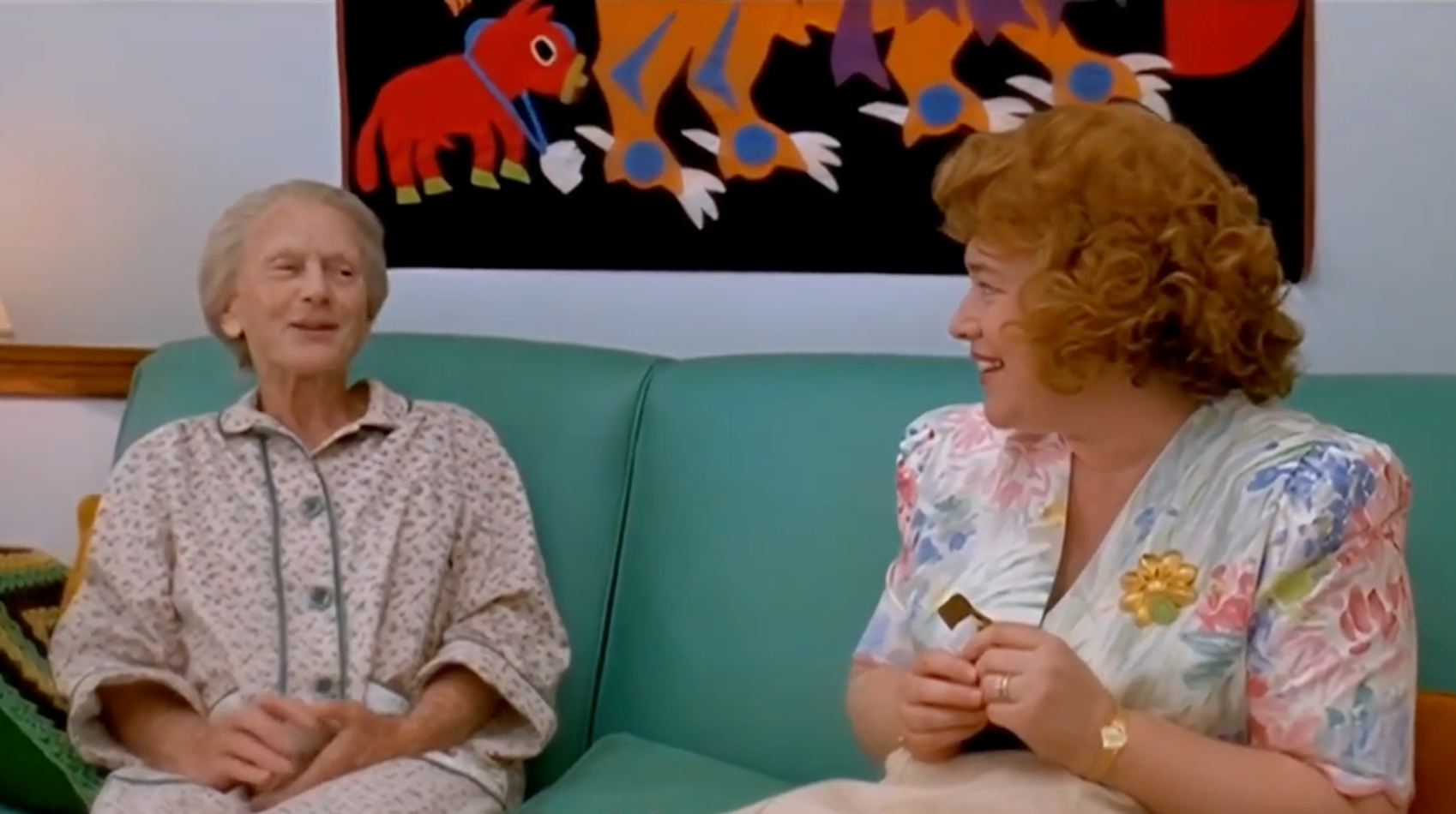 Two people sitting on a couch, one in a floral dress and the other in a patterned robe, conversing and smiling