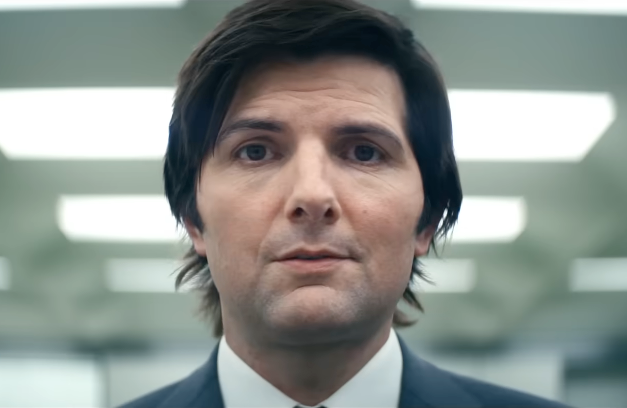 A person with straight hair wearing a suit stands indoors with a neutral expression, in a well-lit environment