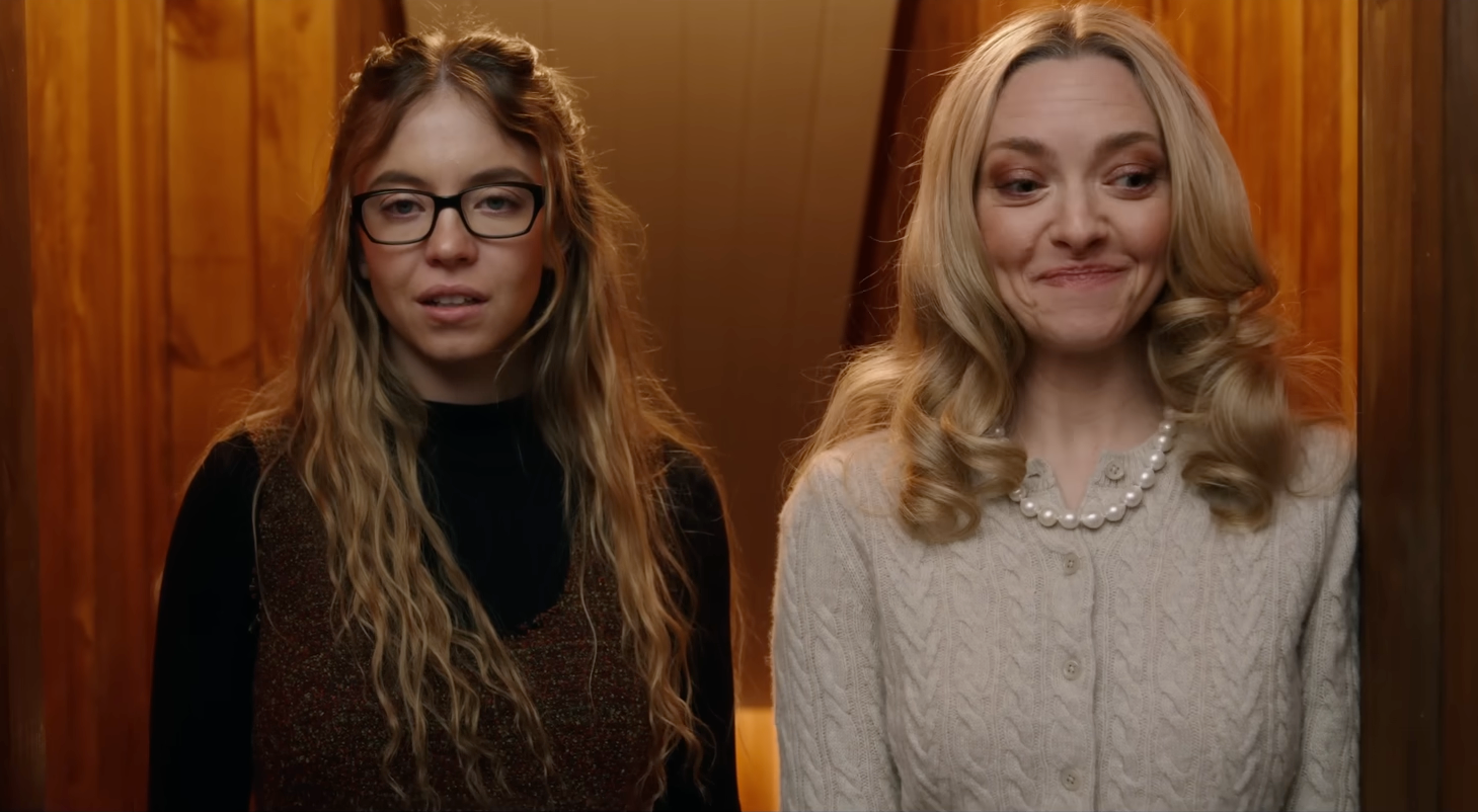 Two women stand side-by-side indoors. One wears glasses and a sweater vest; the other wears a knit sweater with pearl accessories