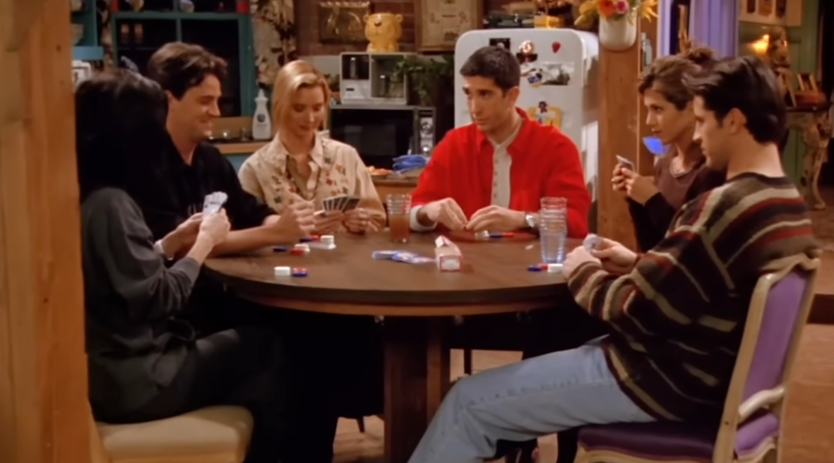 Six people sit around a table playing cards in a cozy room, concentrating on the game