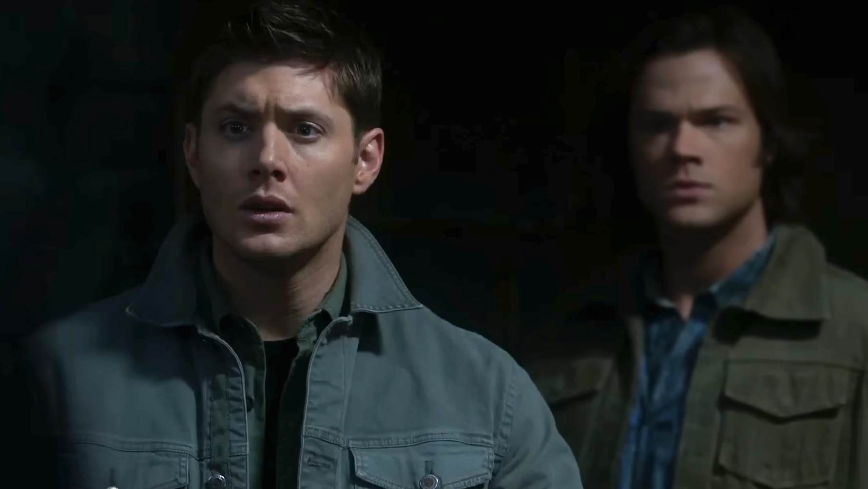 Two men stand in a dimly lit space, looking intently forward. Both wear casual jackets