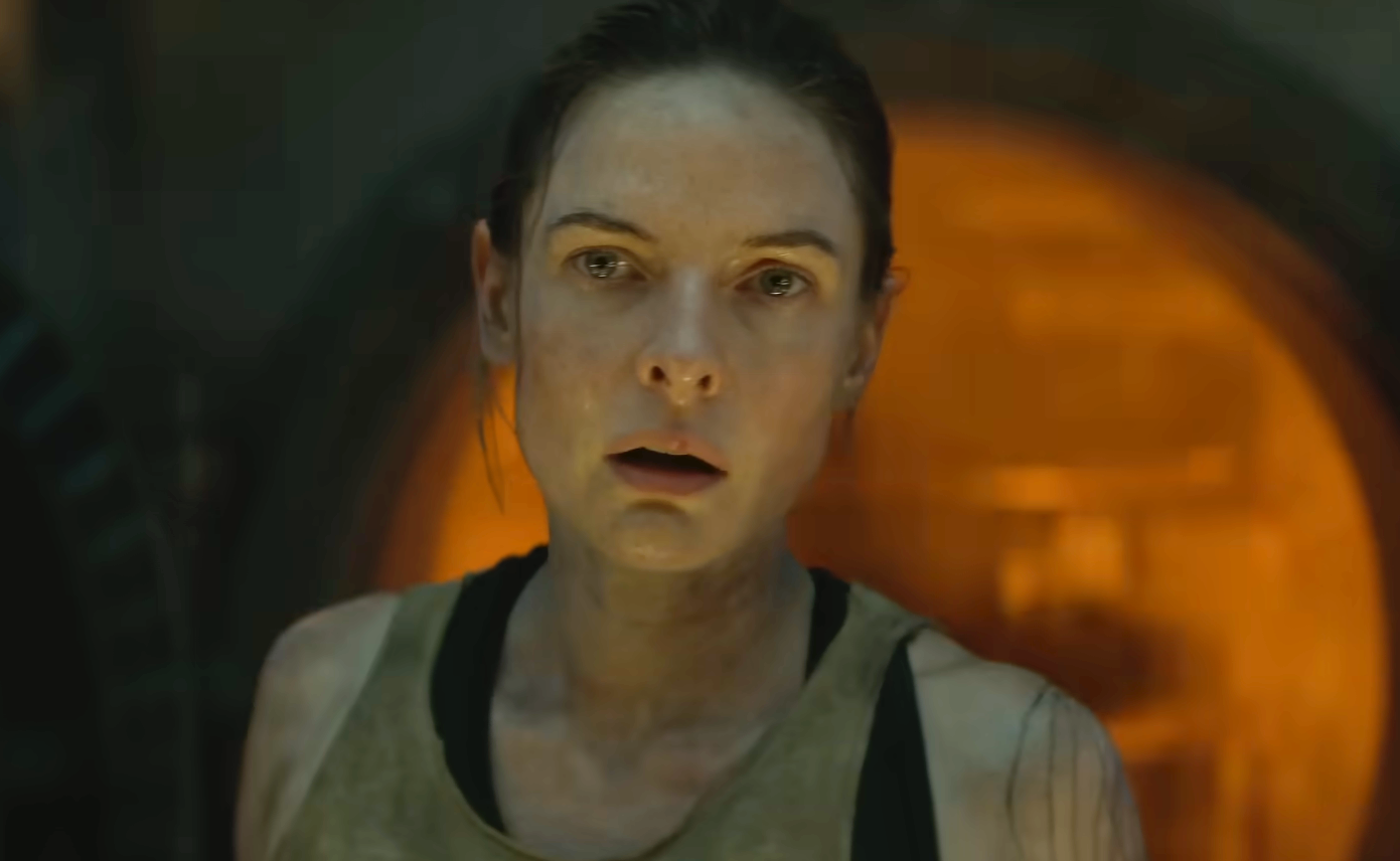 A person with a focused expression, wearing a sleeveless top, stands in front of an orange-lit, industrial background