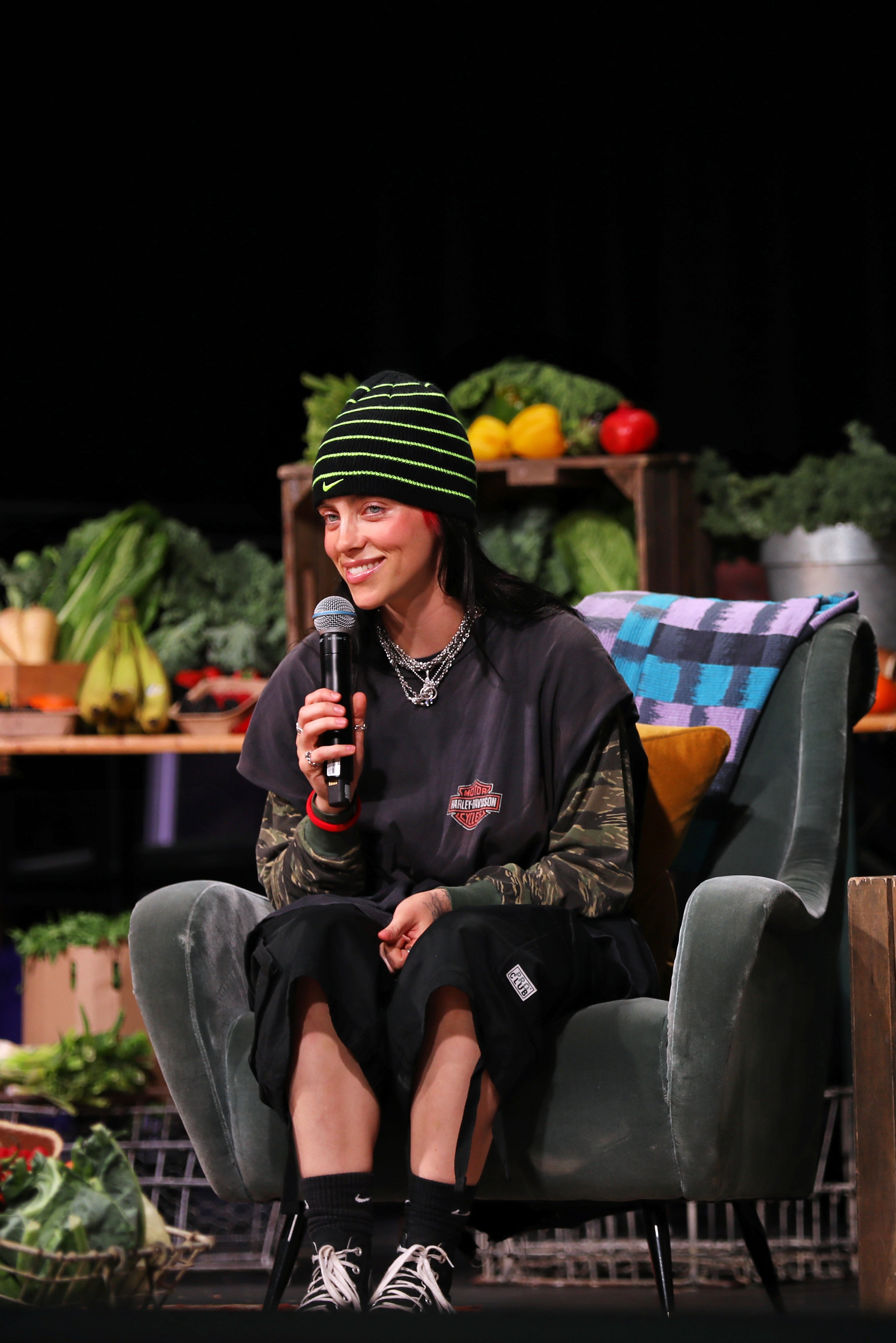 Billie Eilish speaks onstage during Overheated, a one-off climate activism event presented by Support + Feed, surrounded by a vibrant vegetable display in the background