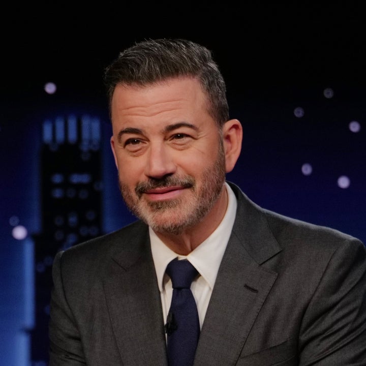 A person in a suit sits at a desk with a nameplate reading "Jimmy Kimmel," in a late-night talk show setting