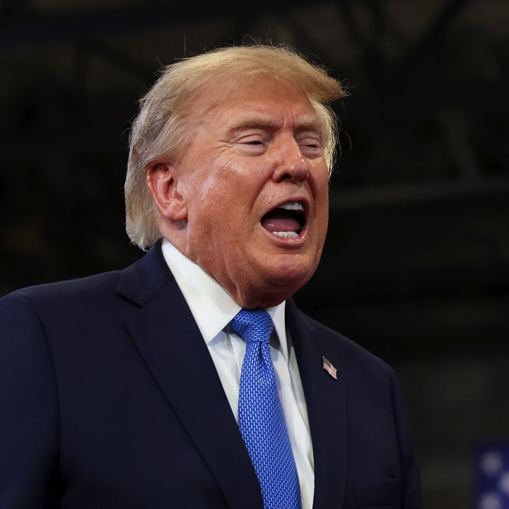 A man in a suit and tie speaks energetically at an event, with a large American flag in the background