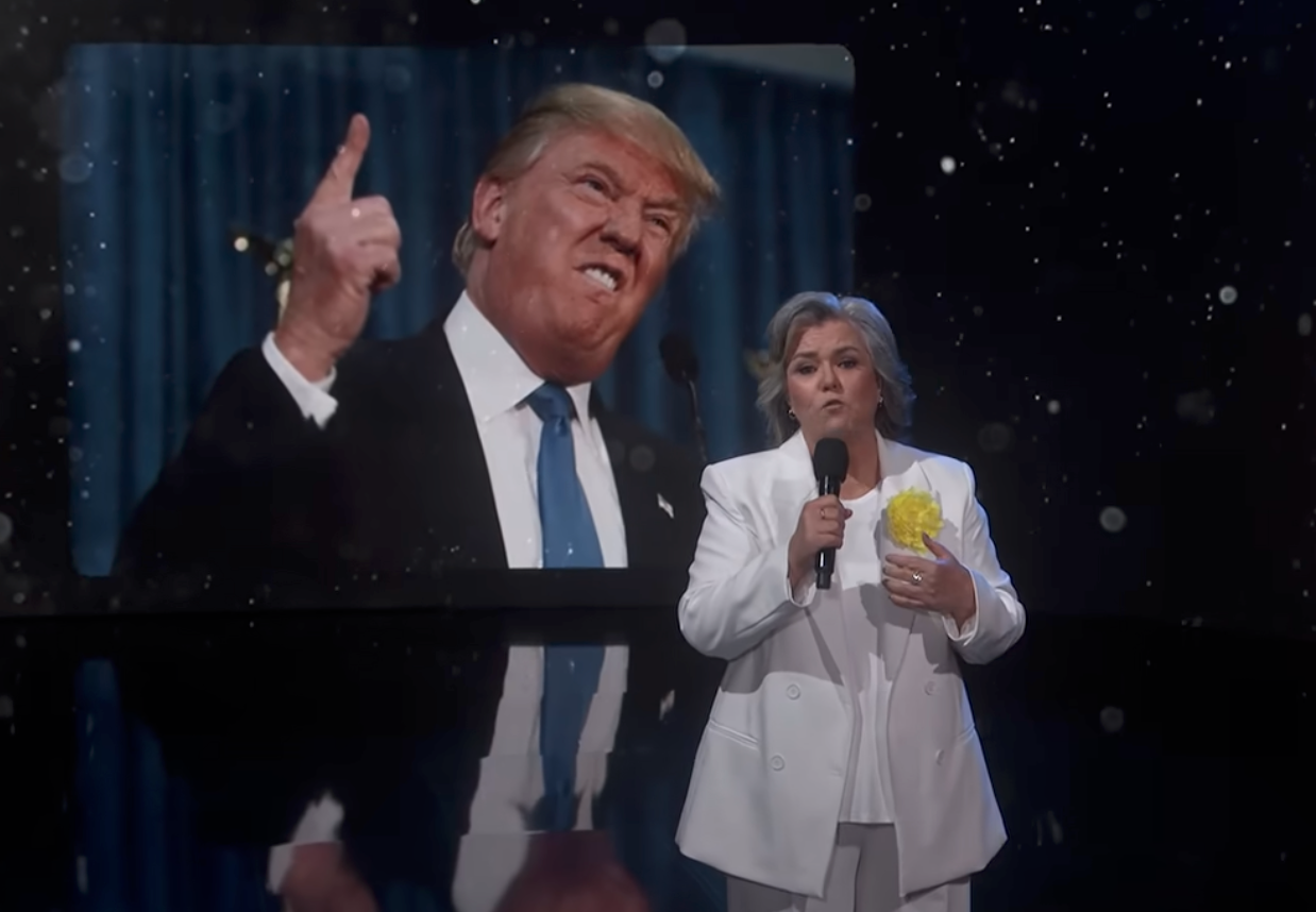 A person in a white suit delivers a comedic monologue on stage, with an image of a political figure gesturing in the background