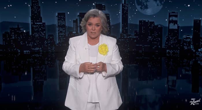 Person in a white suit with a yellow flower, standing in front of a cityscape backdrop, possibly during a TV show or event