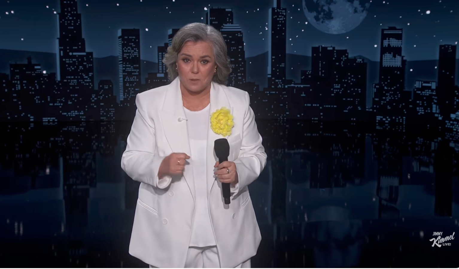 A person wearing a white suit with a yellow flower brooch holds a microphone on a nighttime cityscape stage