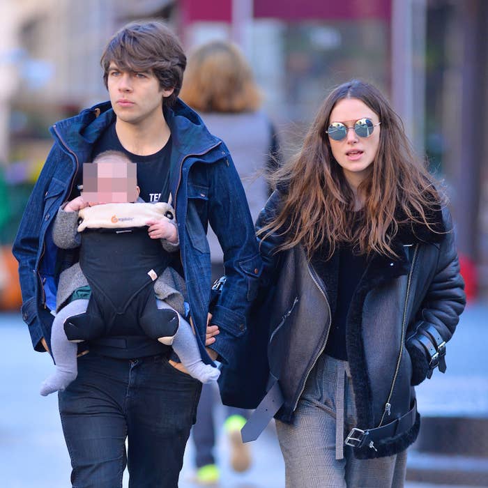 Two people walking with a baby in a carrier. One wears a denim jacket, and the other wears a leather jacket with sunglasses