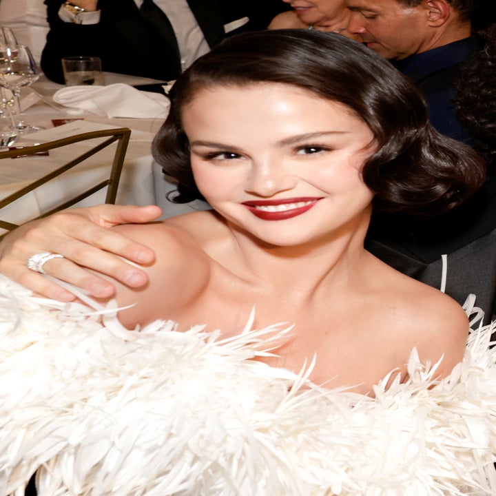 Group of people seated at a formal event. One person in a feathered outfit, others in suits, smiling at the table with glasses and decorations