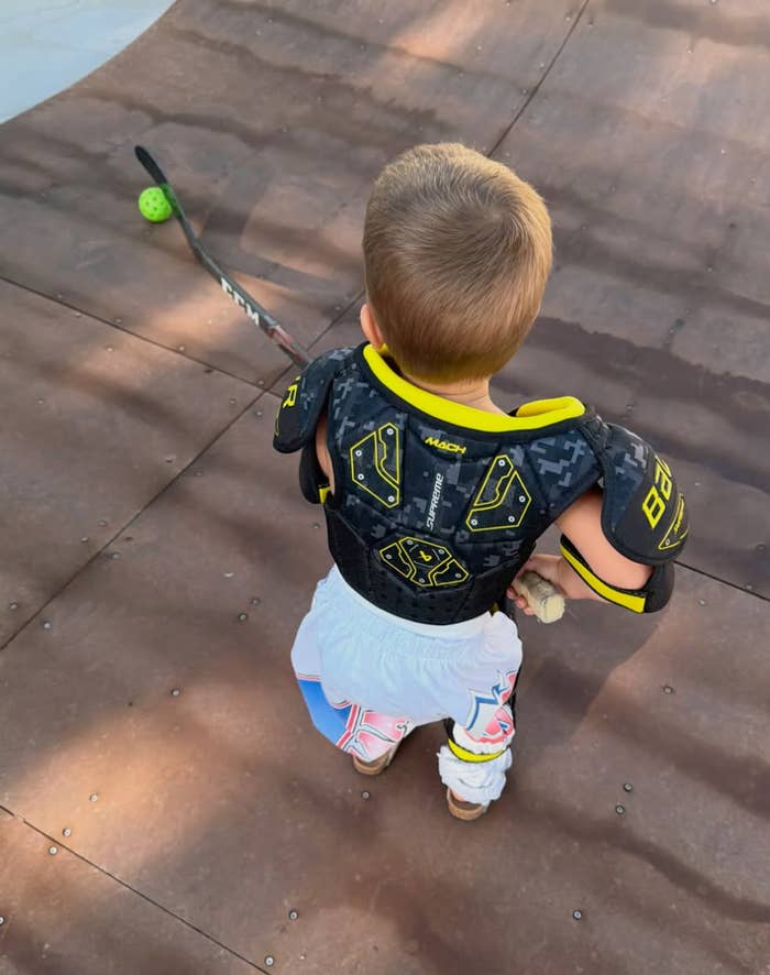 Child in sports gear kneels on a skate ramp with a hockey stick, looking towards a green ball