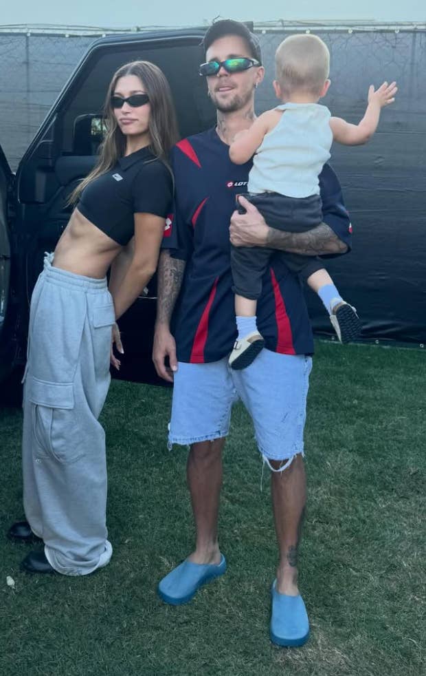 A woman in a crop top and cargo pants poses with a man in casual sportswear holding a toddler, outdoors