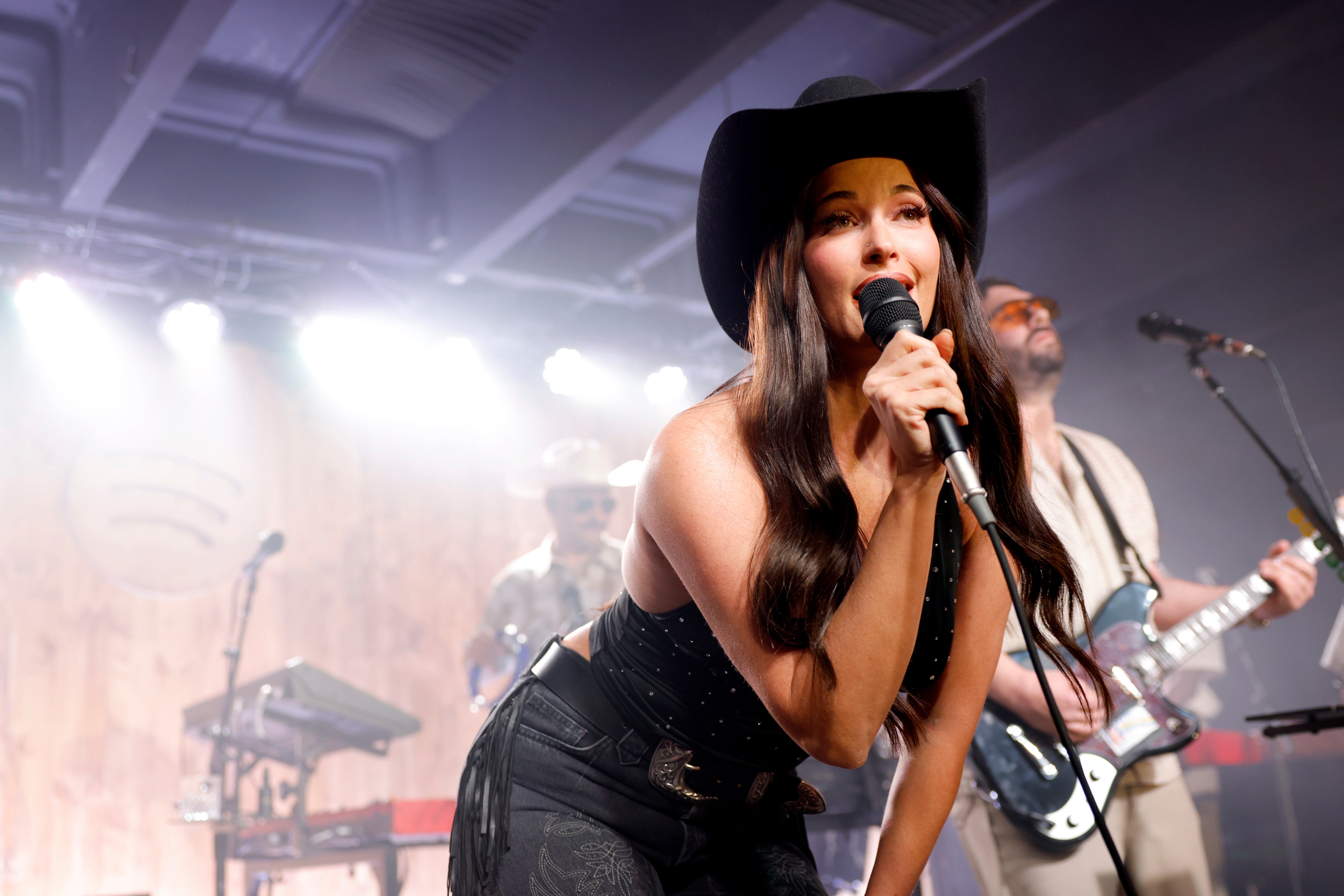 Sanger i cowboyhatt opptrer energisk på scenen, holder en mikrofon, med et band som spiller instrumenter i bakgrunnen
