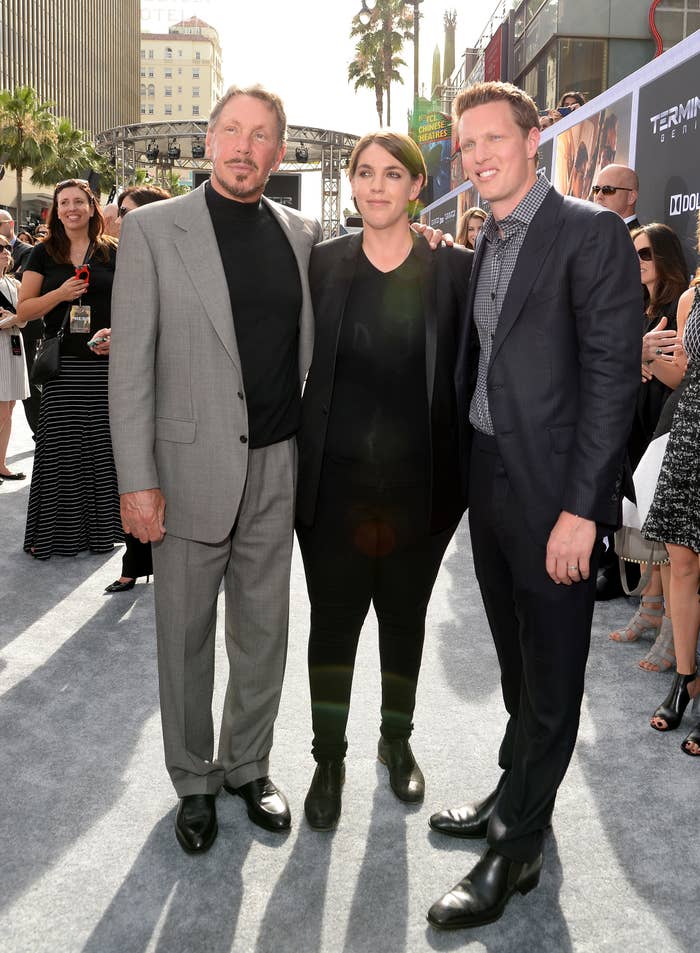 Three people pose on a red carpet. One wears a suit, another a black outfit with a jacket, and the third a suit with a checked shirt