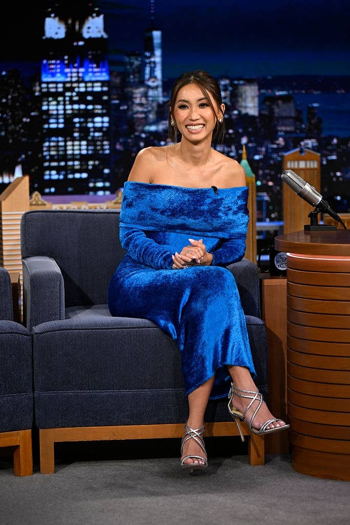 Person seated on a talk show set, wearing an off-shoulder textured dress and strappy heels, smiling. Cityscape backdrop visible