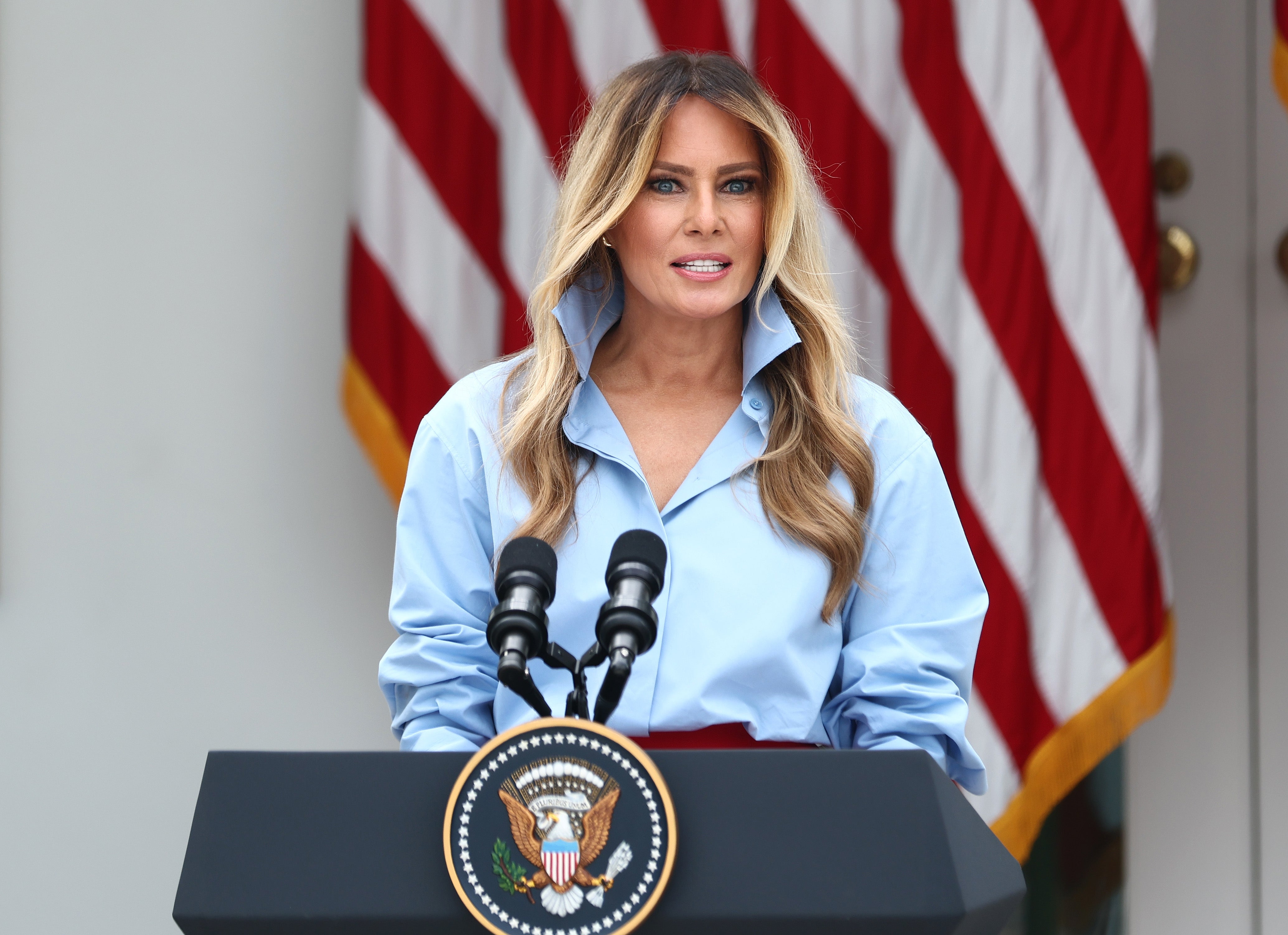 En kvinne holder en tale utendørs på et podium med den amerikanske presidentens segl, iført en stilig skjorte med krage med amerikanske flagg i bakgrunnen