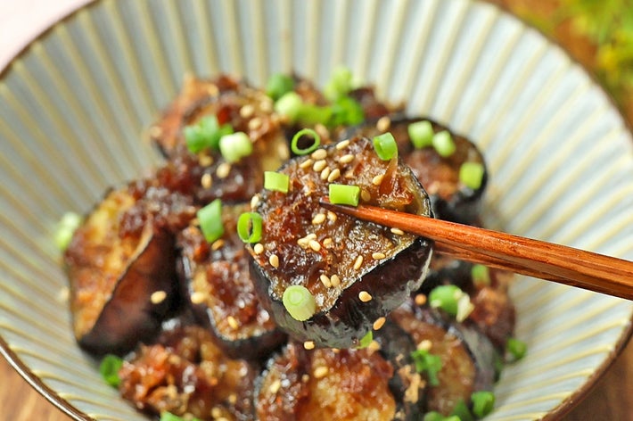 皿に盛られた煮込みなすの上に、小ねぎとゴマがトッピングされている料理。箸で一切れを持ち上げている。
