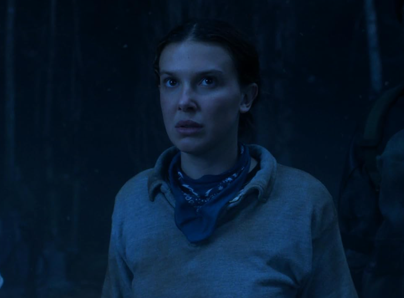Person in a tense scene, wearing a scarf and casual top, looking determined amidst a dimly lit background