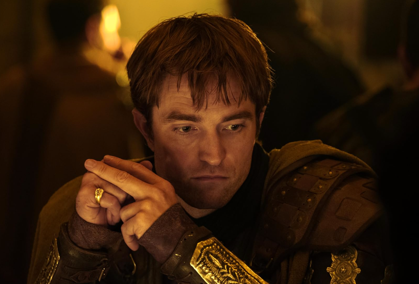 Person in medieval armor, seated, with chin resting on hands. Wearing a ring, looking pensive. Indoor setting with soft lighting