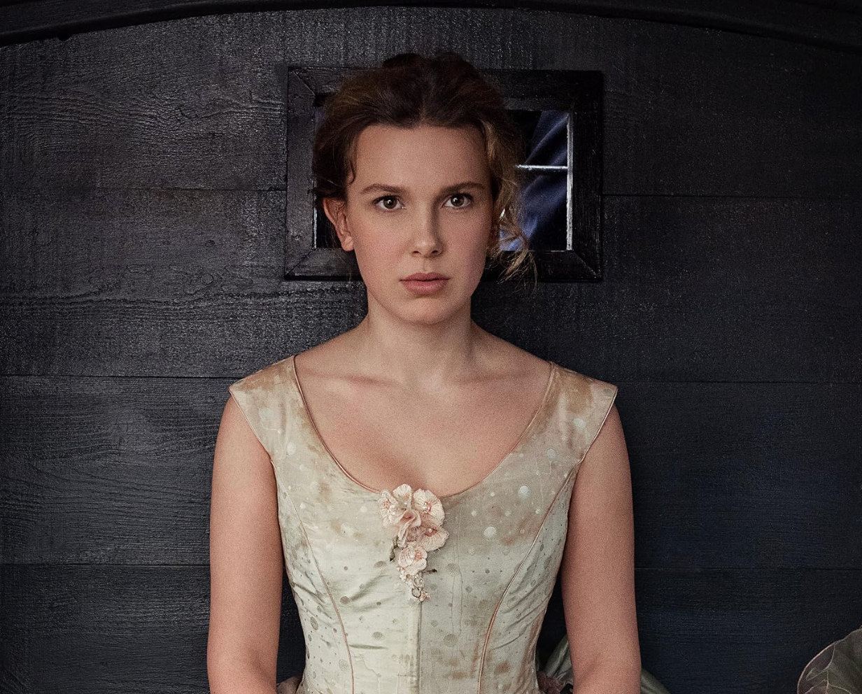 Person in vintage-style dress with floral details, standing against a dark wooden background, looking directly at the camera