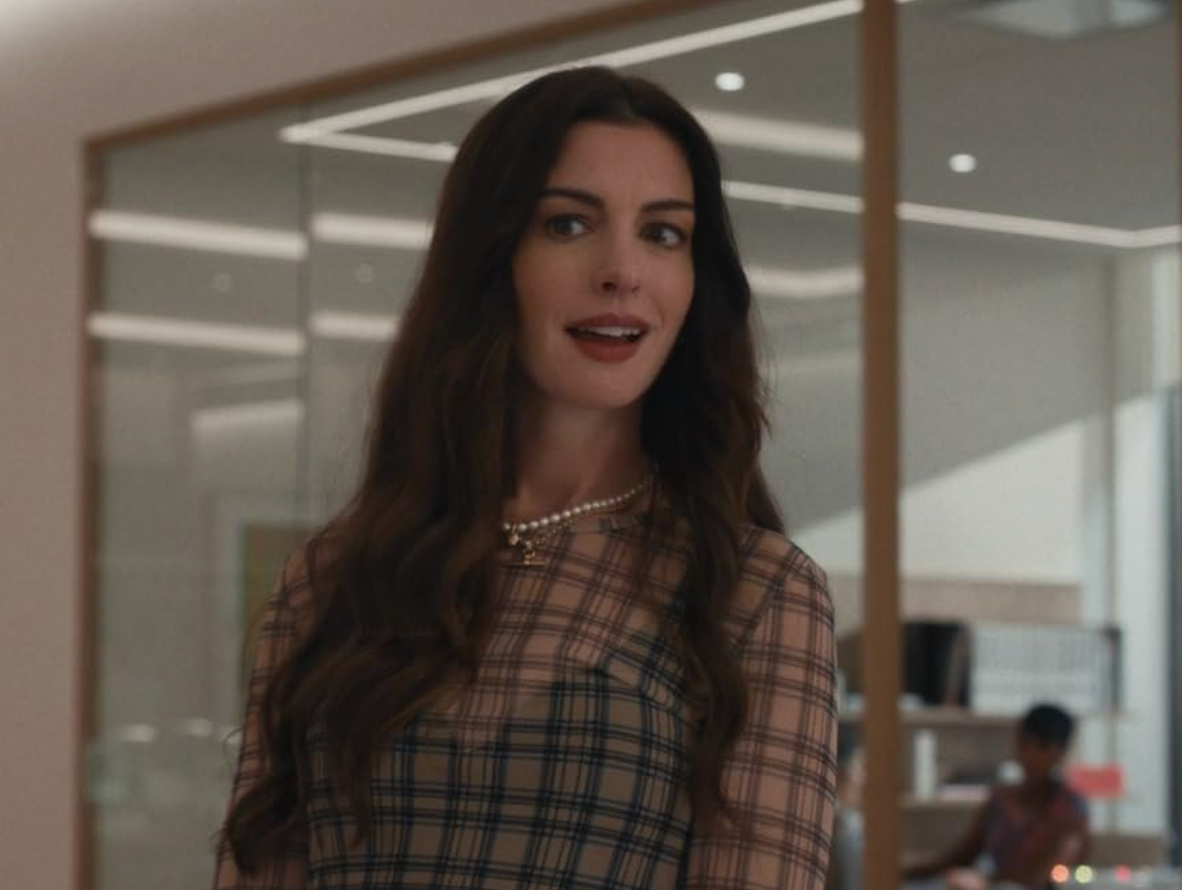 A woman with long hair, wearing a plaid dress and pearl necklace, smiles in a modern office environment